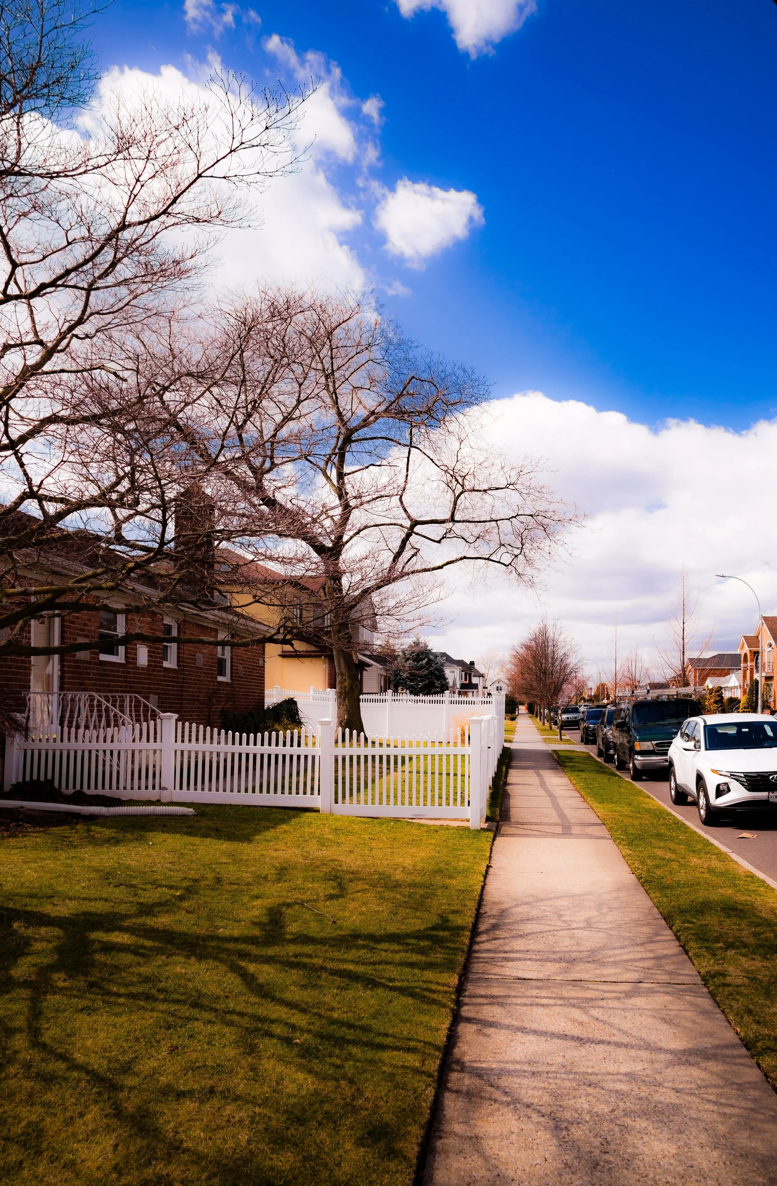 Residential neighborhood on a clear day with a sidewalk, parked cars, a white picket fence, leafless trees, and a blue sky with clouds.