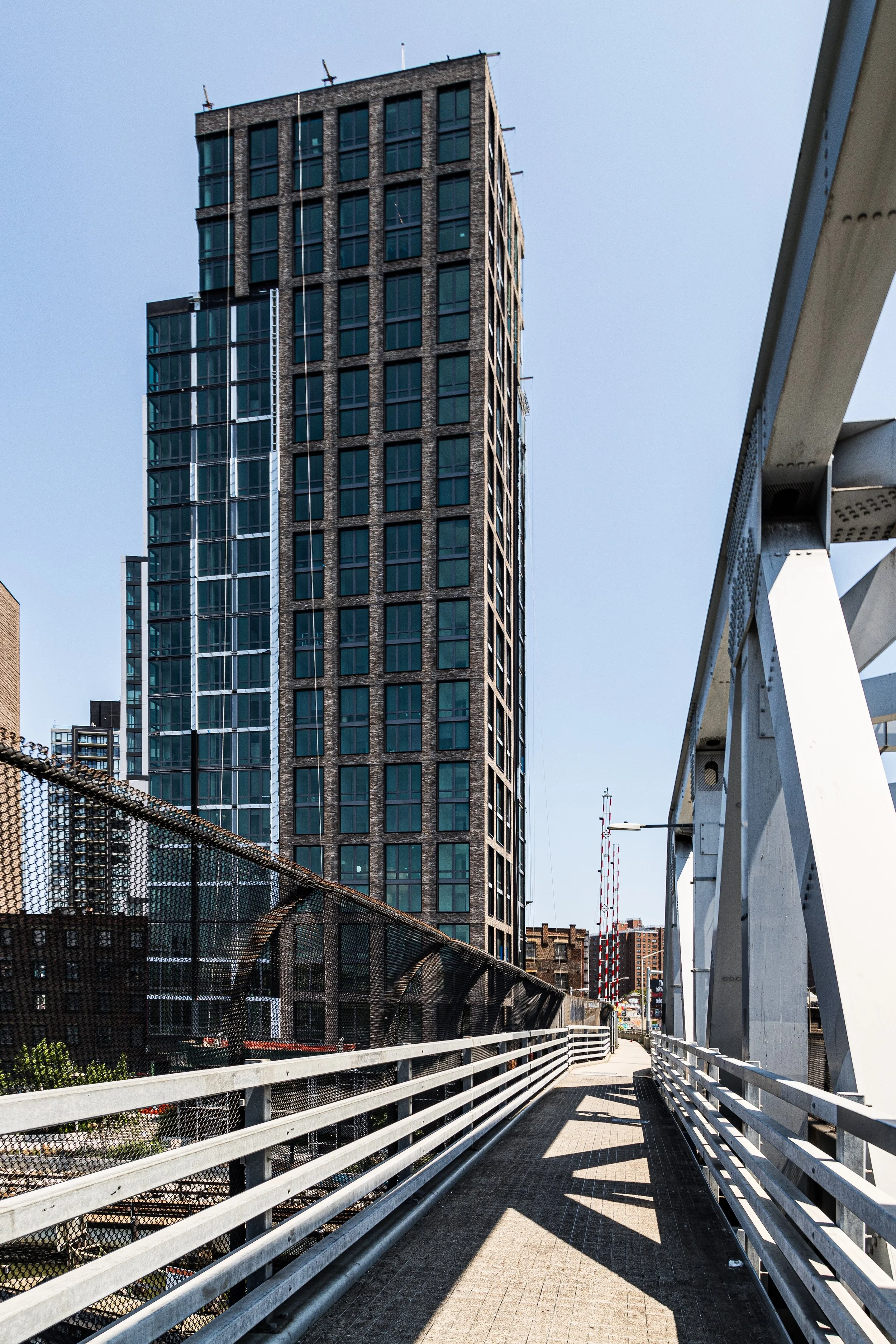 A high-rise building viewed from a pedestrian bridge with metal railings and a white support structure, with a clear sky in the background.