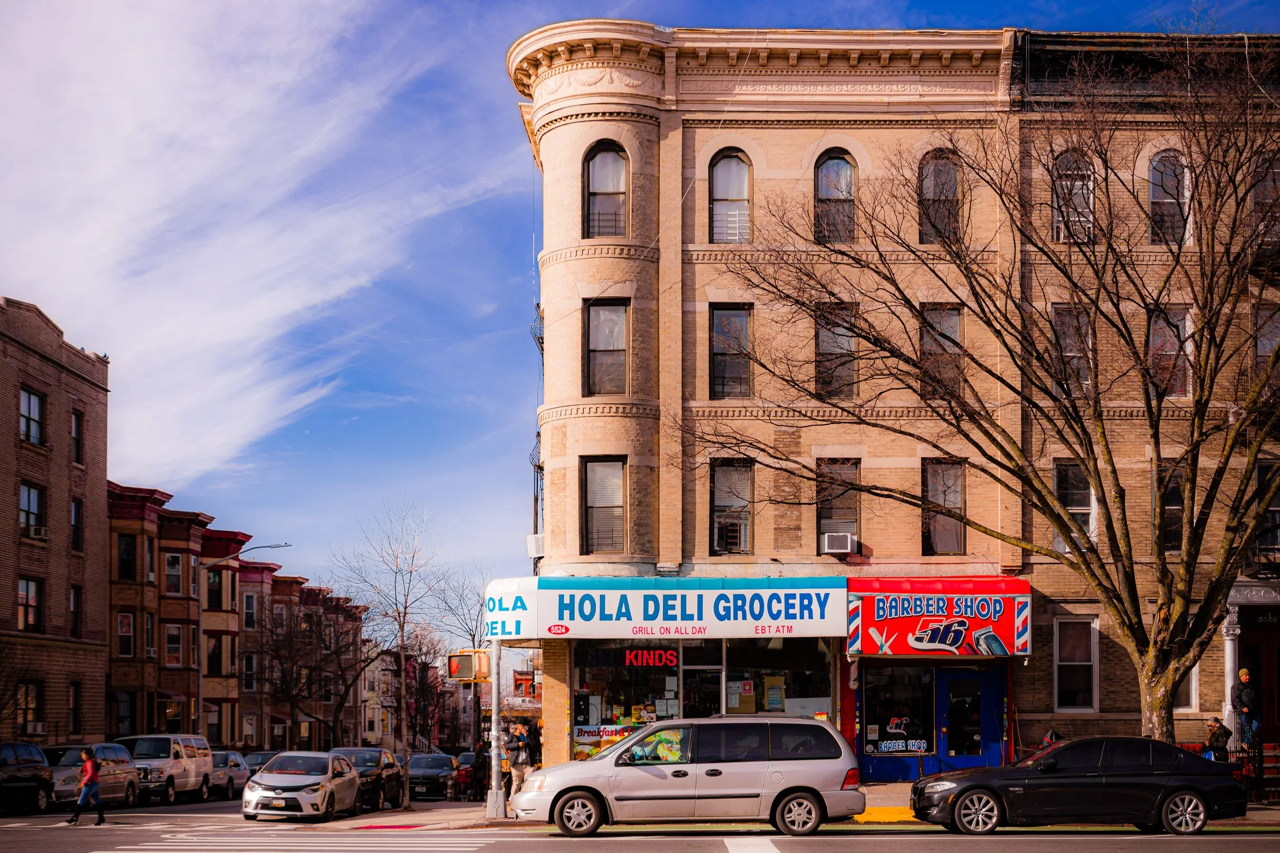 Street view of a corner building with a grocery store named 'Hola Deli Grocery' and a barber shop. Several parked cars and pedestrians are visible on the street. The building is made of beige bricks and has a rounded corner with multiple windows. A leafless tree is in front of the building.