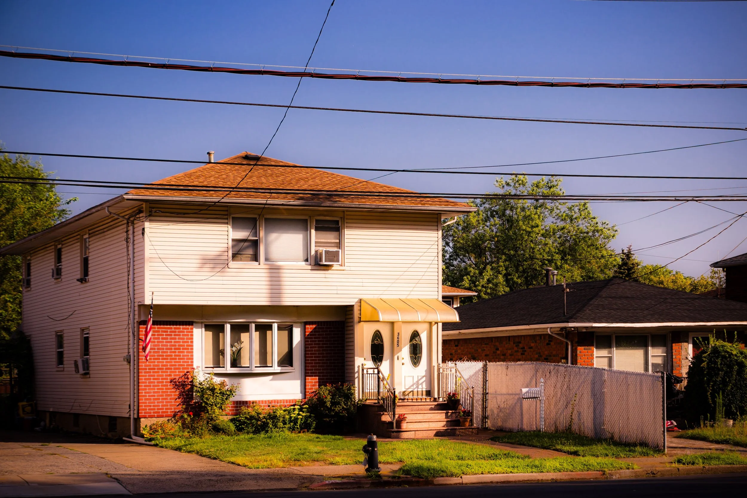 A two-story house with beige siding, brick accents, and a brown tiled roof. There are three windows on the top floor, one of which has an air conditioning unit. The front door has an oval glass design and is accessed via steps. There are plants and bushes in front of the house, a sidewalk, and a street with a fire hydrant. Power lines cross the sky above the house.