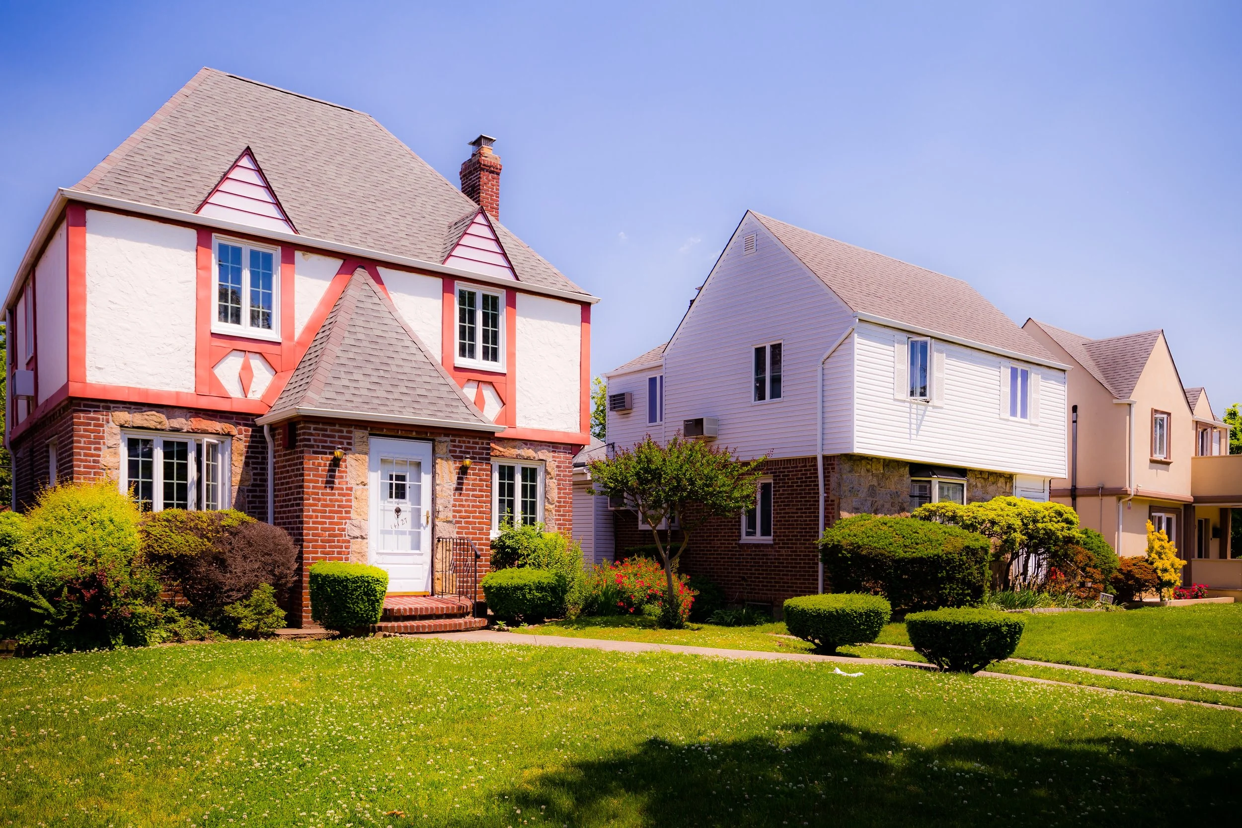 Front view of three houses with well-maintained lawns, small bushes, and trees under a clear blue sky.