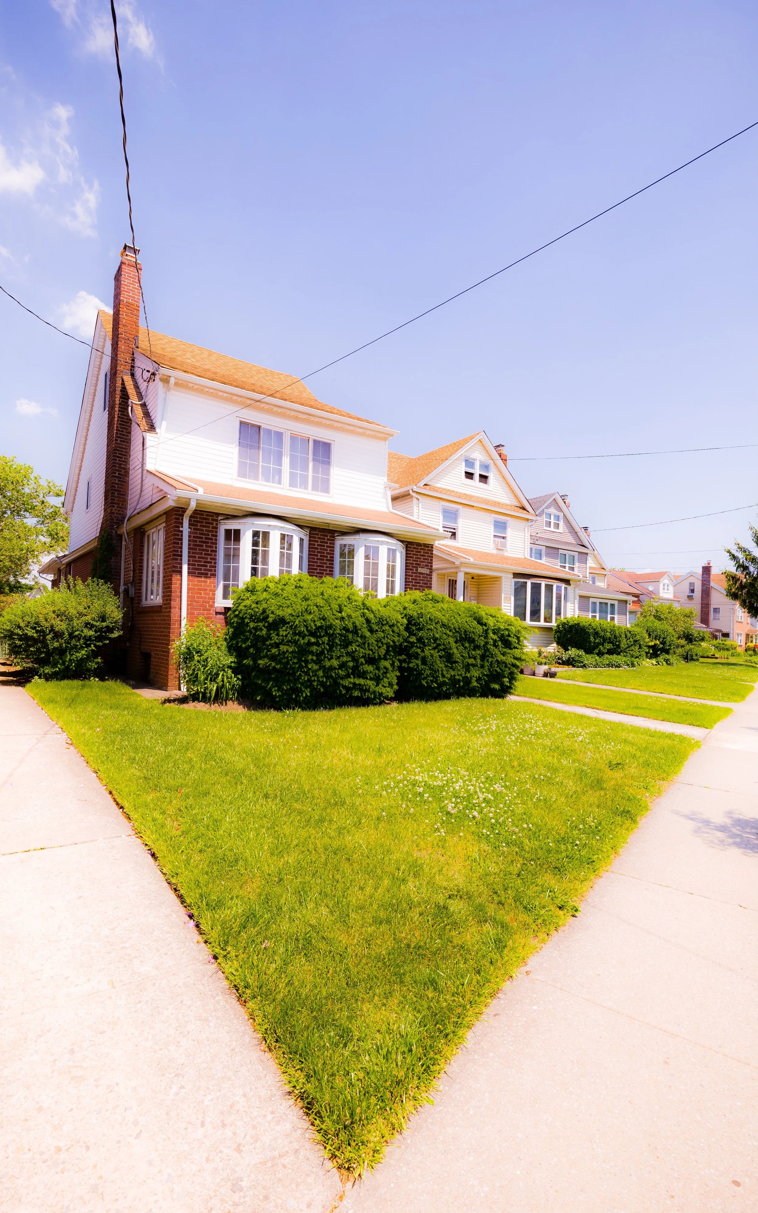 A row of Victorian-style houses with brick and white siding, surrounded by lush green lawns and bushes, under a clear blue sky.