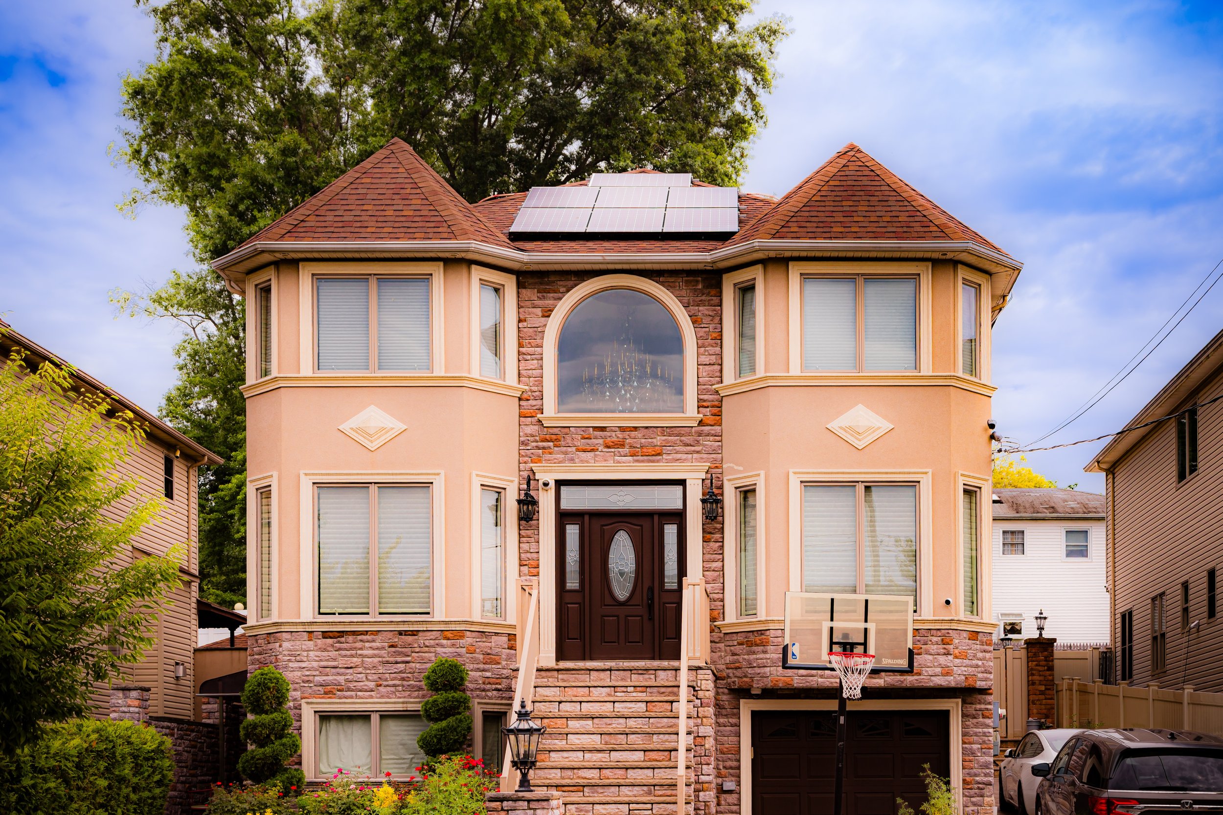 A large, two-story house with a brick and beige stucco exterior, curved bay windows, and a central arched window on the upper level. The house has a dark front door, solar panels on the roof, and a small set of stairs leading to the entrance. Two trees and some shrubs are in the front yard, and there is a basketball hoop in the driveway.