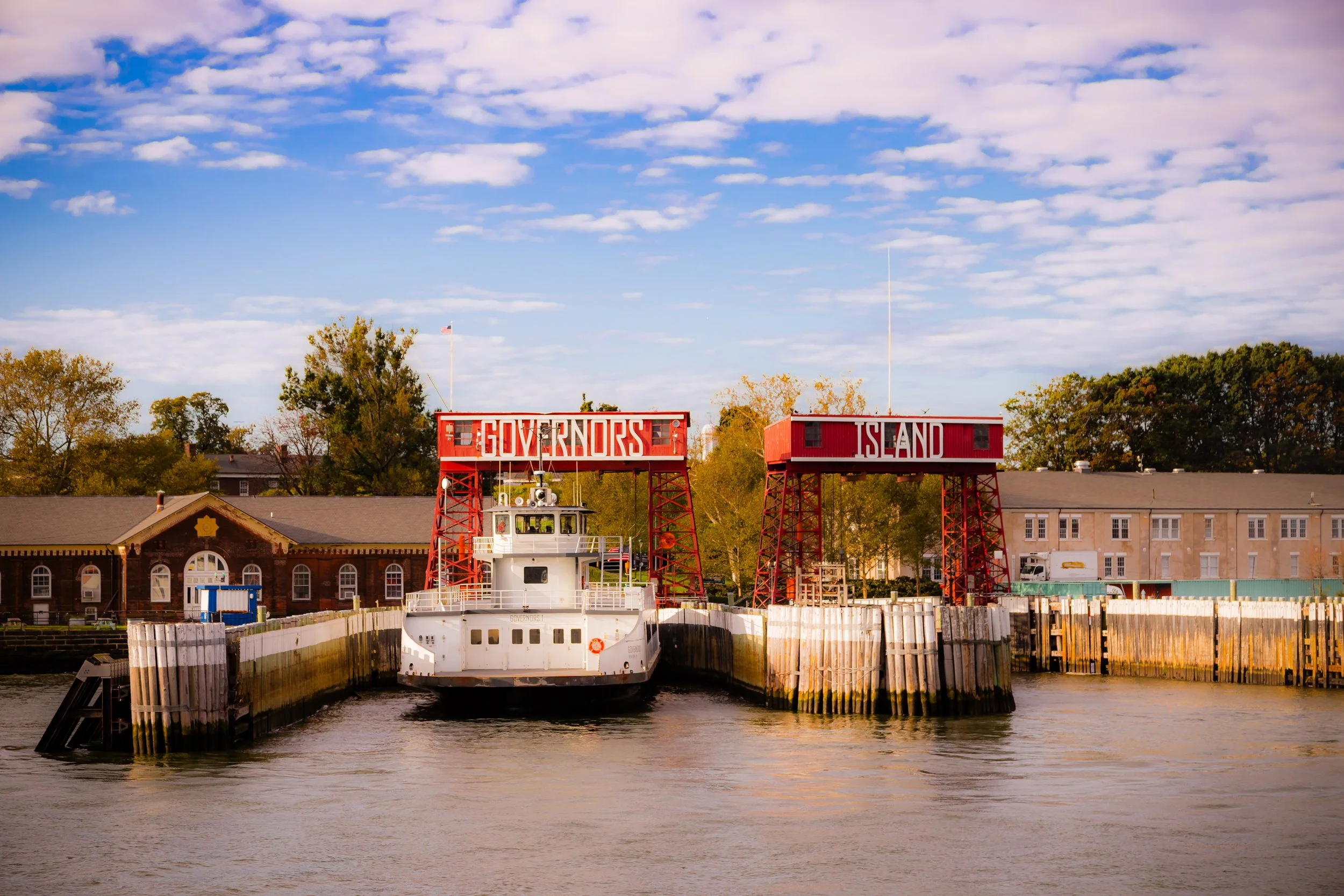 Ferry dock with a boat passing through a canal, red signs reading 'GOVERNORS' and 'ISLAND' on metal structures, trees and buildings in the background, blue sky with clouds.