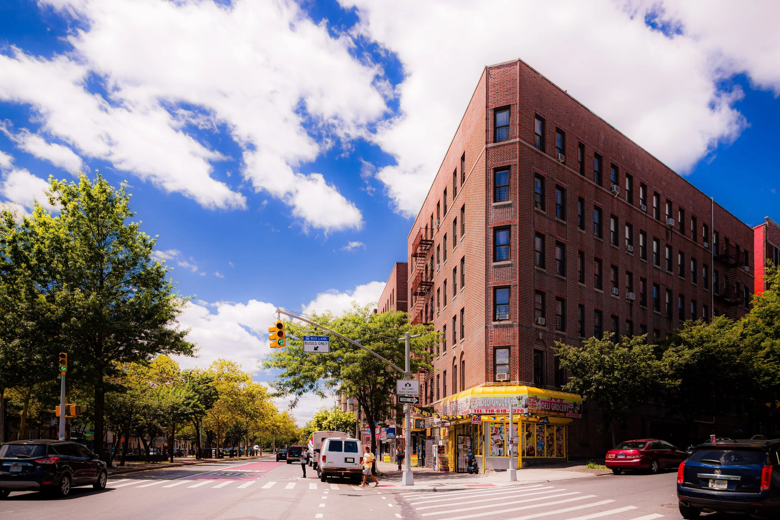 A city street scene featuring a large brick building on a sunny day with white clouds in the sky, trees lining the sidewalk, cars and pedestrians crossing the intersection, and a small grocery store with a yellow canopy.