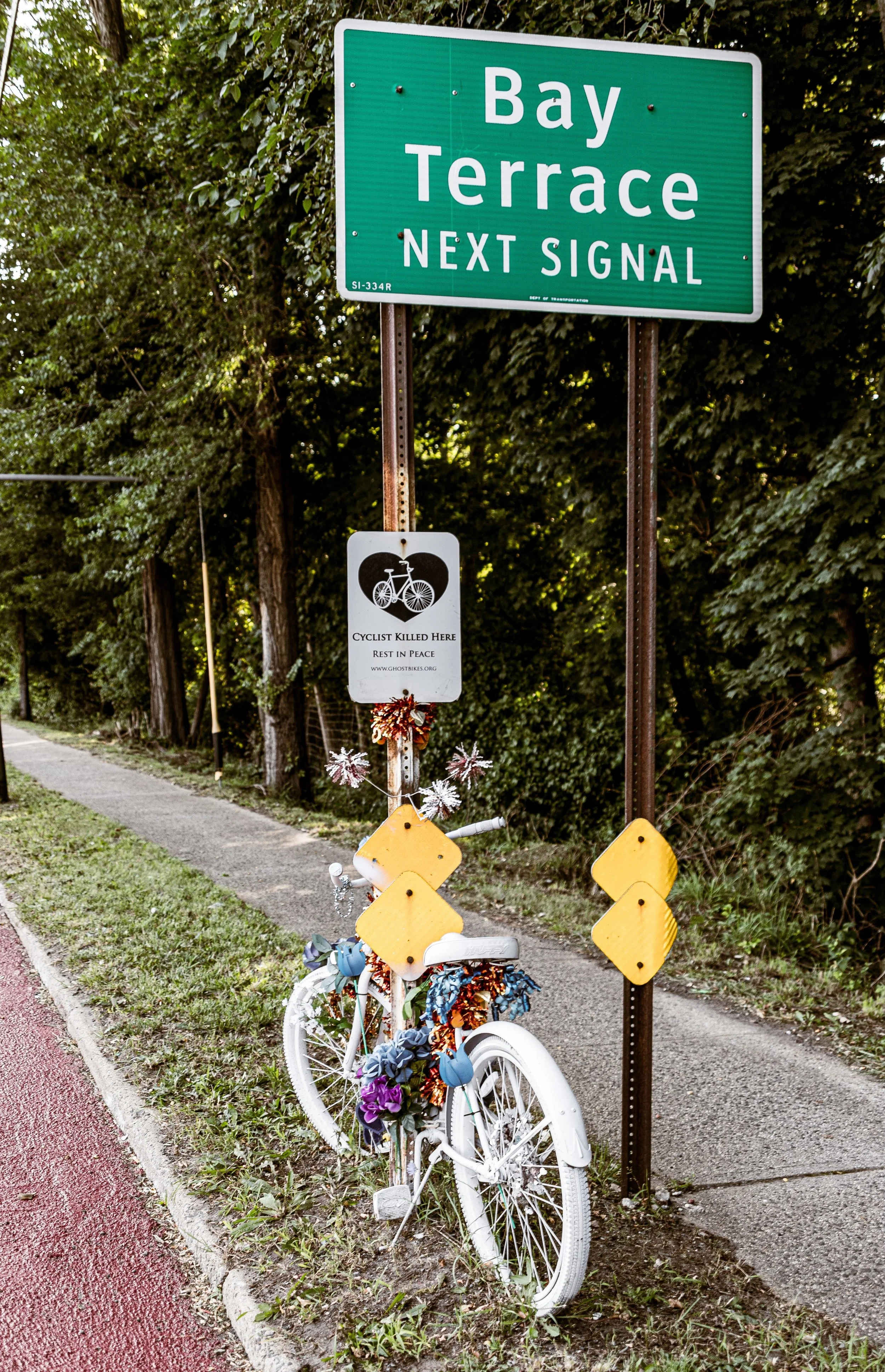 A green street sign that says 'Bay Terrace Next Signal' with a smaller sign below referencing a cyclist memorial. A decorated white bicycle is positioned next to the signs.
