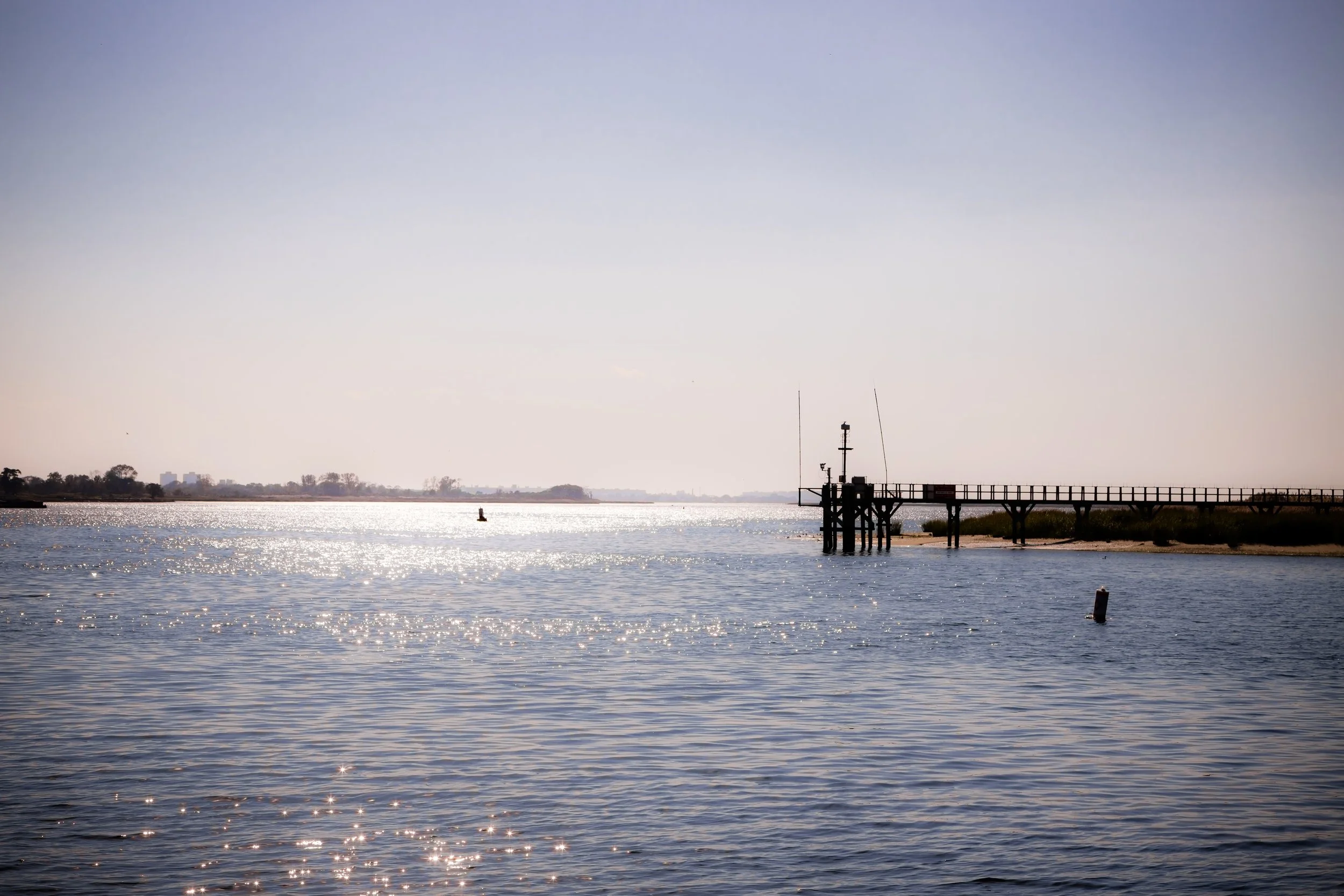 View of a calm body of water with sunlight reflecting off the surface. A wooden pier extends into the water on the right side of the image. In the background, there is a distant shoreline with some trees and a clear sky.