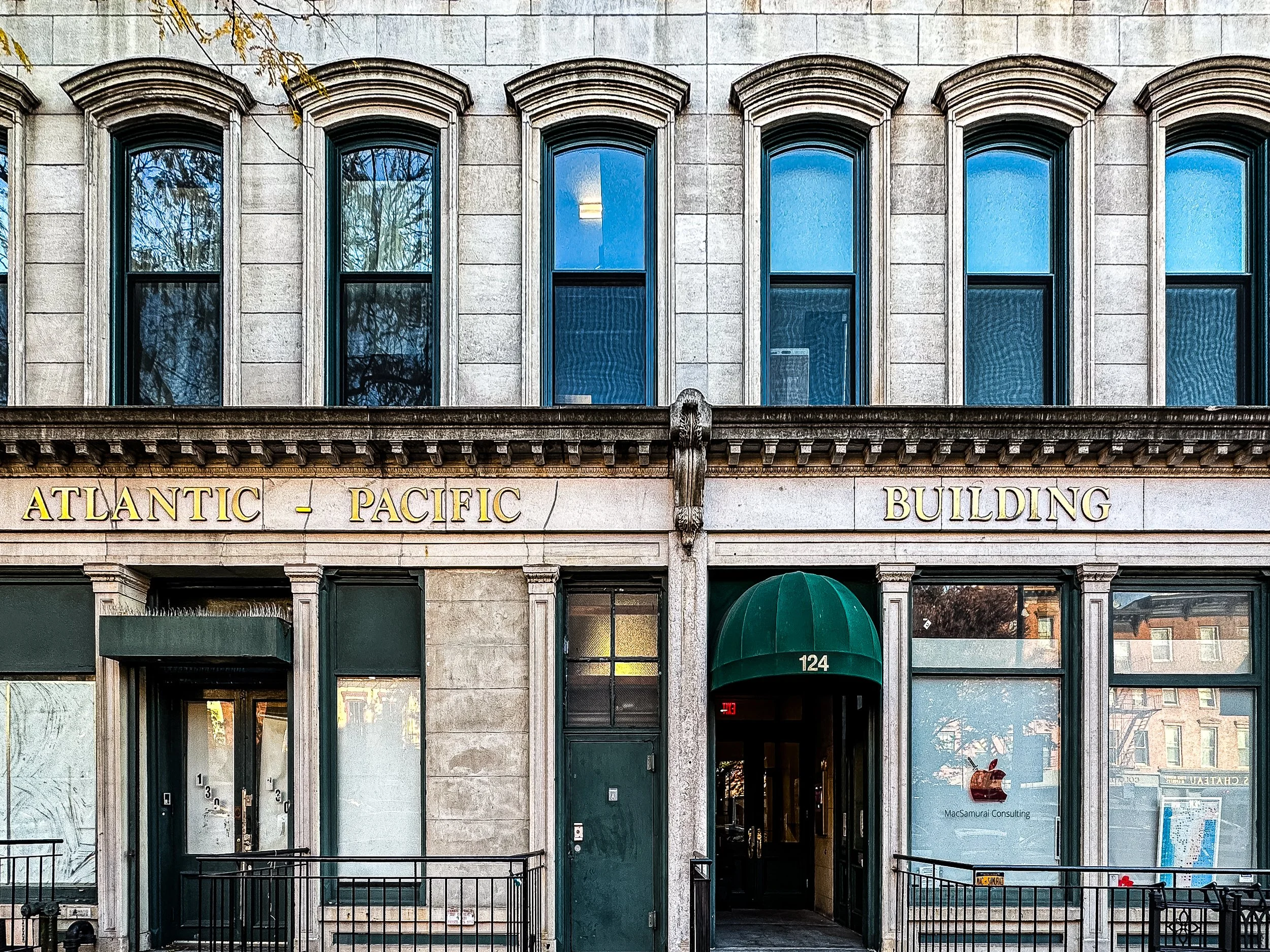 Exterior of an old brick building with signs reading 'Atlantic - Pacific Building'. There is a green awning over the entrance, which is numbered 124, leading inside. Large windows reflect nearby buildings and trees.