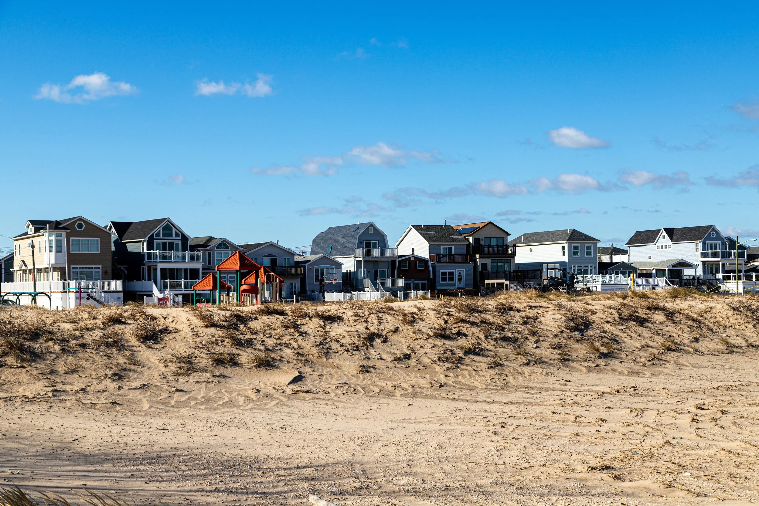 Row of beachfront houses with a sandy beach and grassy dunes in the foreground, under a bright blue sky with a few clouds.
