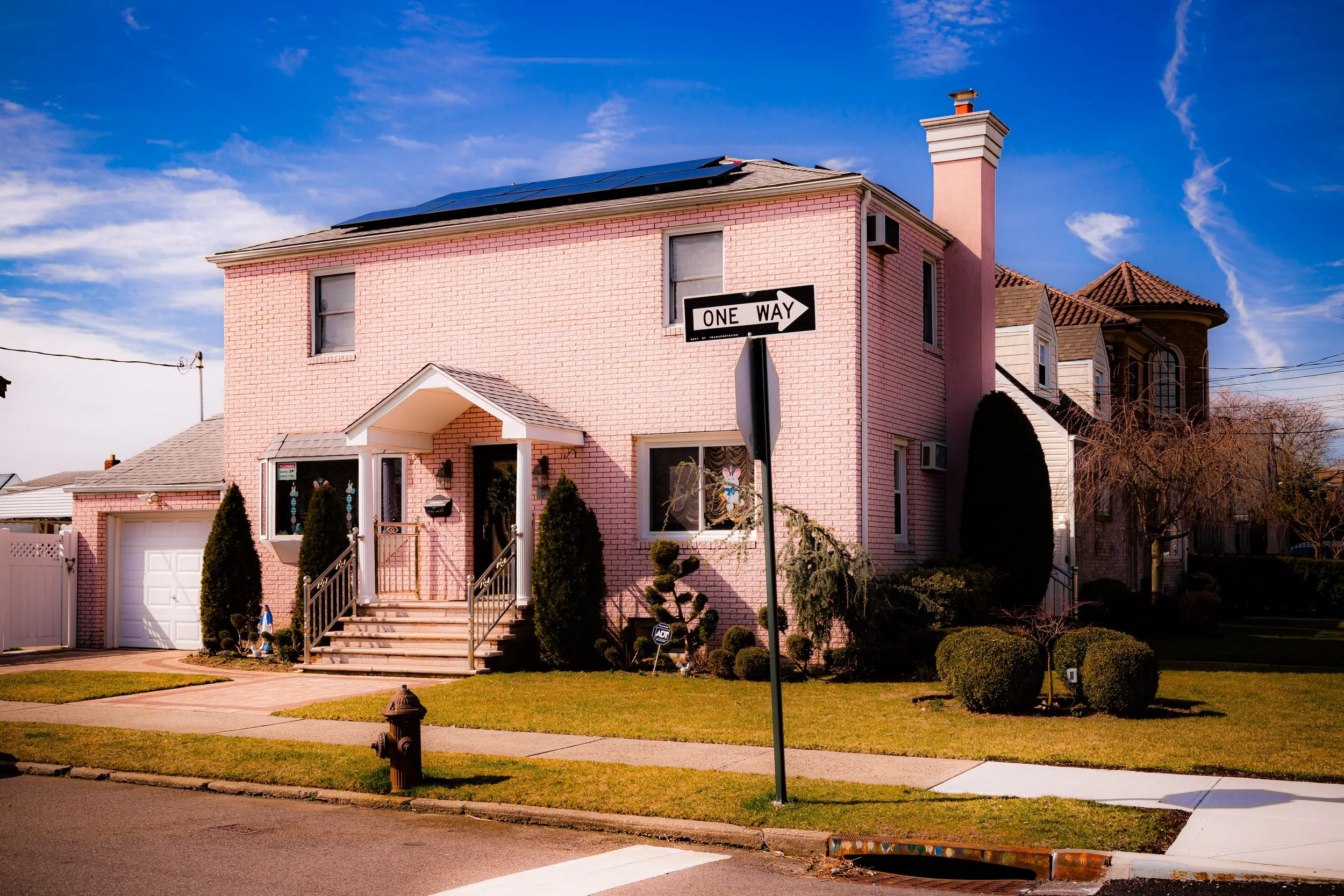 Pink brick house on a corner lot with a white garage, front steps, and a small front yard with trimmed bushes, a fire hydrant, and a street sign that reads 'One Way' against a blue sky with clouds.