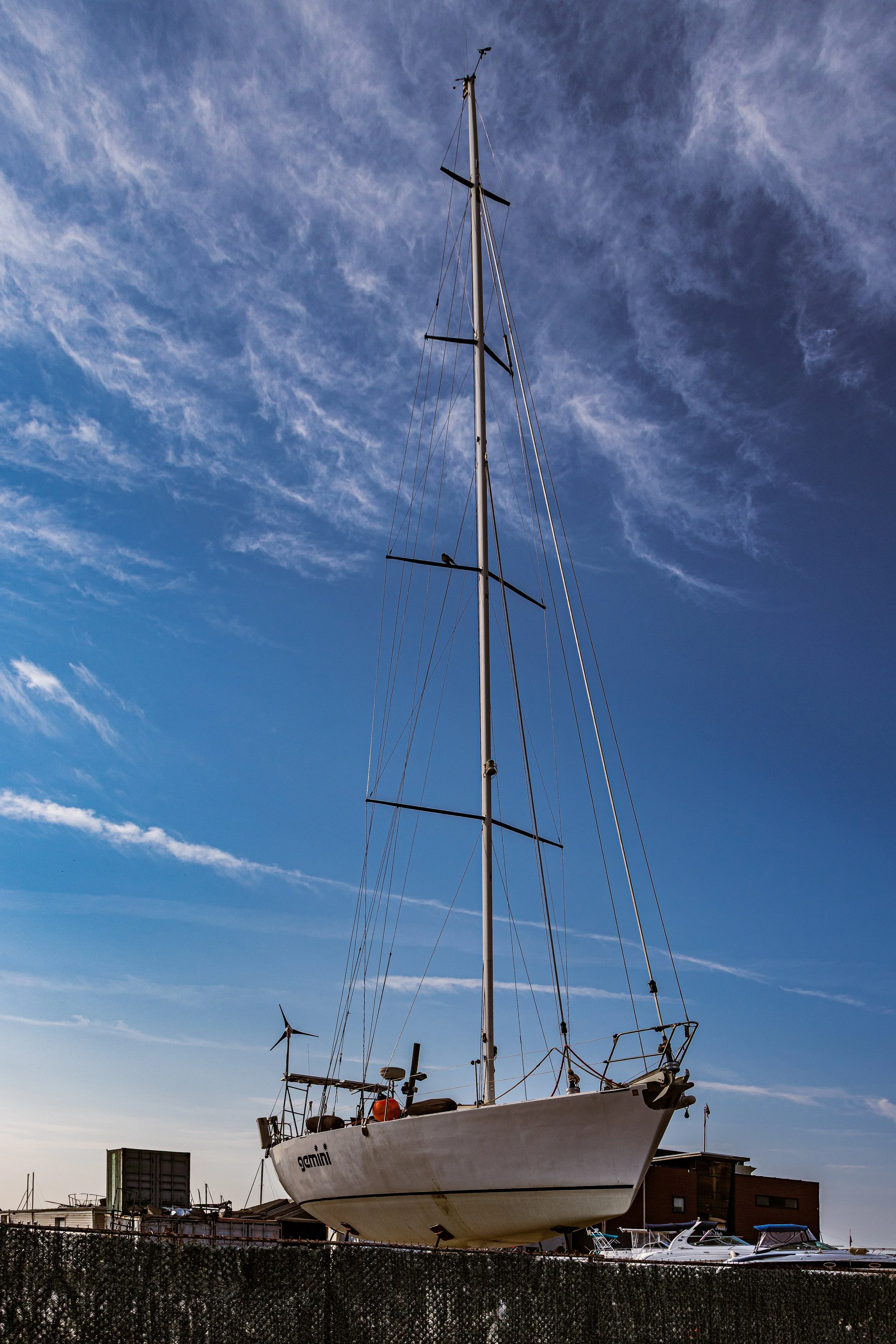 A sailboat named Gamini on land against a blue sky with wispy clouds, nearby buildings and other boats.