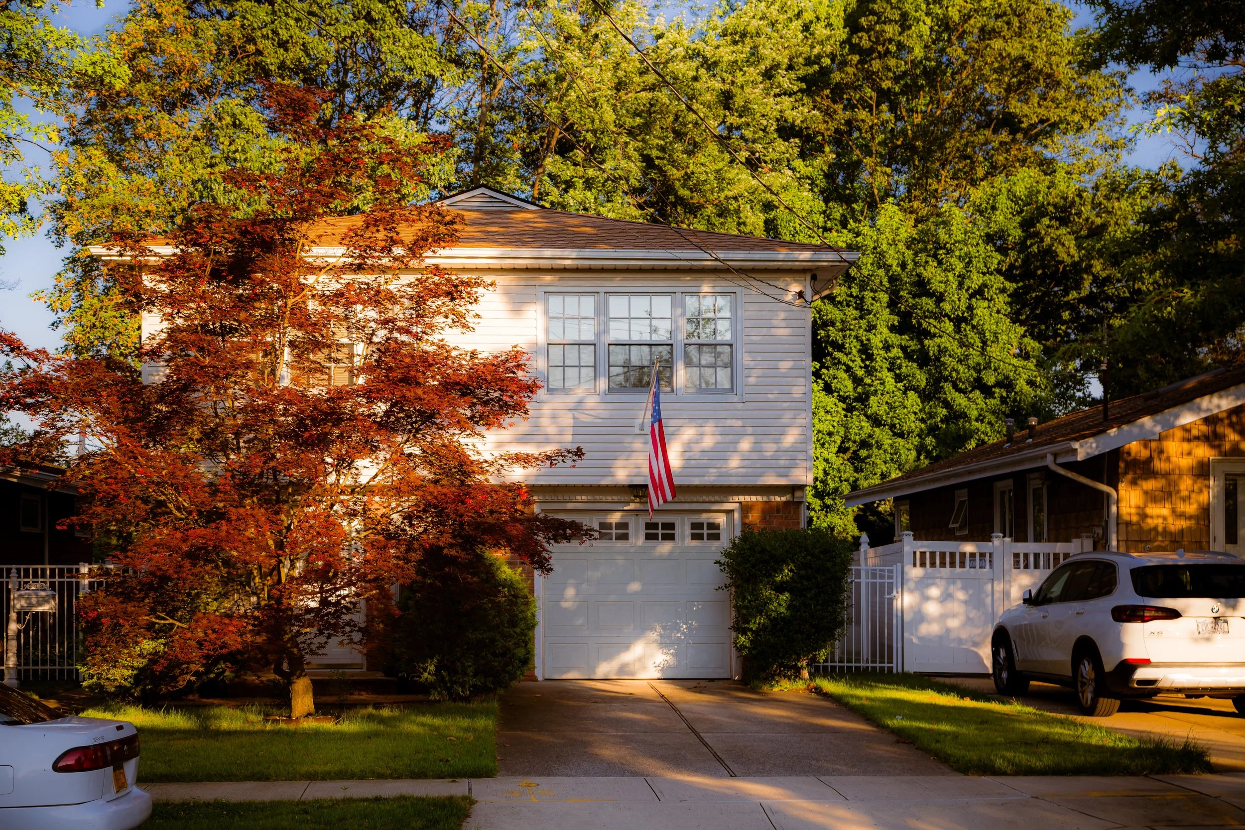 A white two-story house with a garage door, an American flag hanging from a pole near the front window, and a red-leaved tree in front. A white SUV is parked on the driveway, and there are trees and neighboring houses along the street.