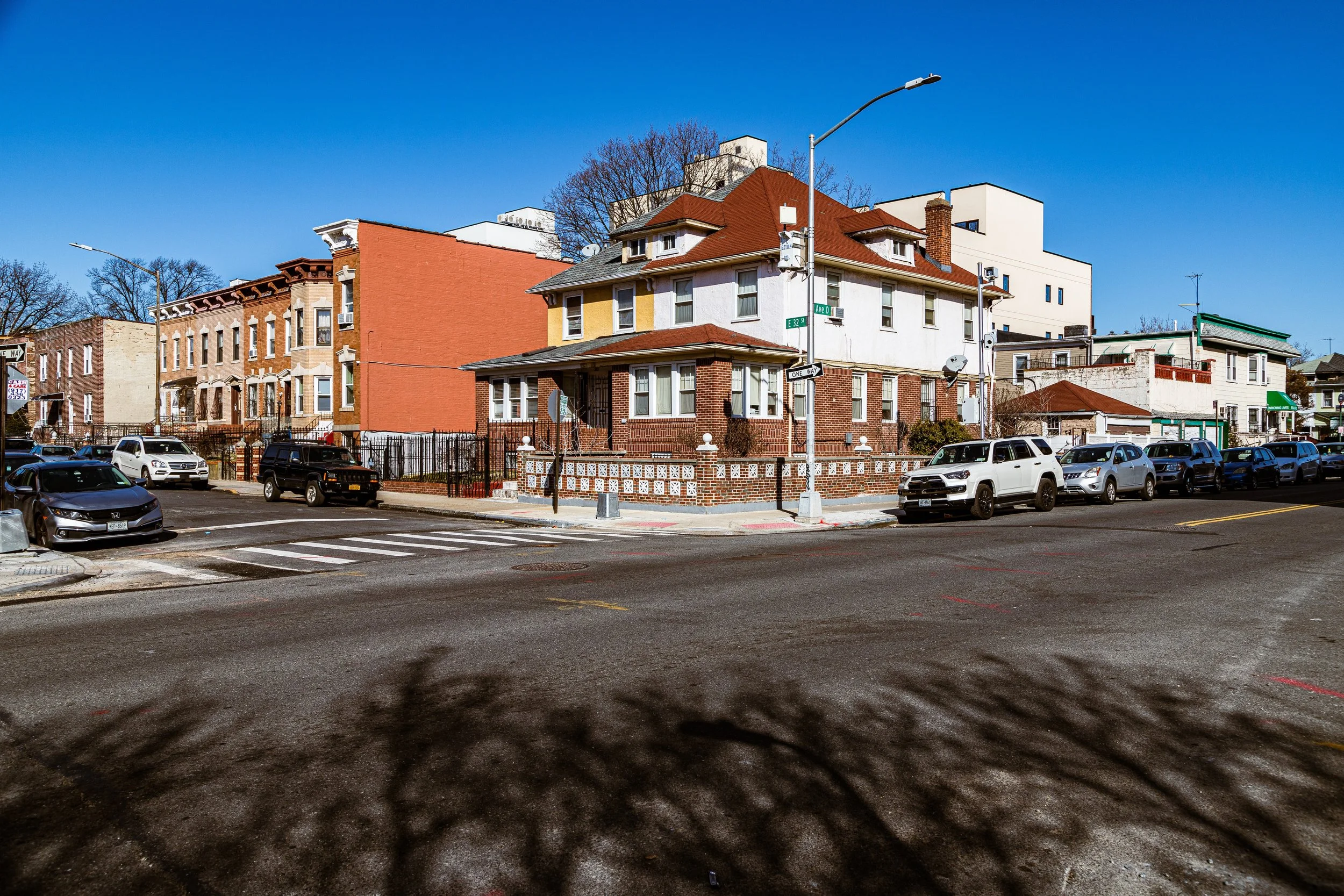 Photo of a street corner with parked cars, a multi-story house at the intersection, and clear blue skies.