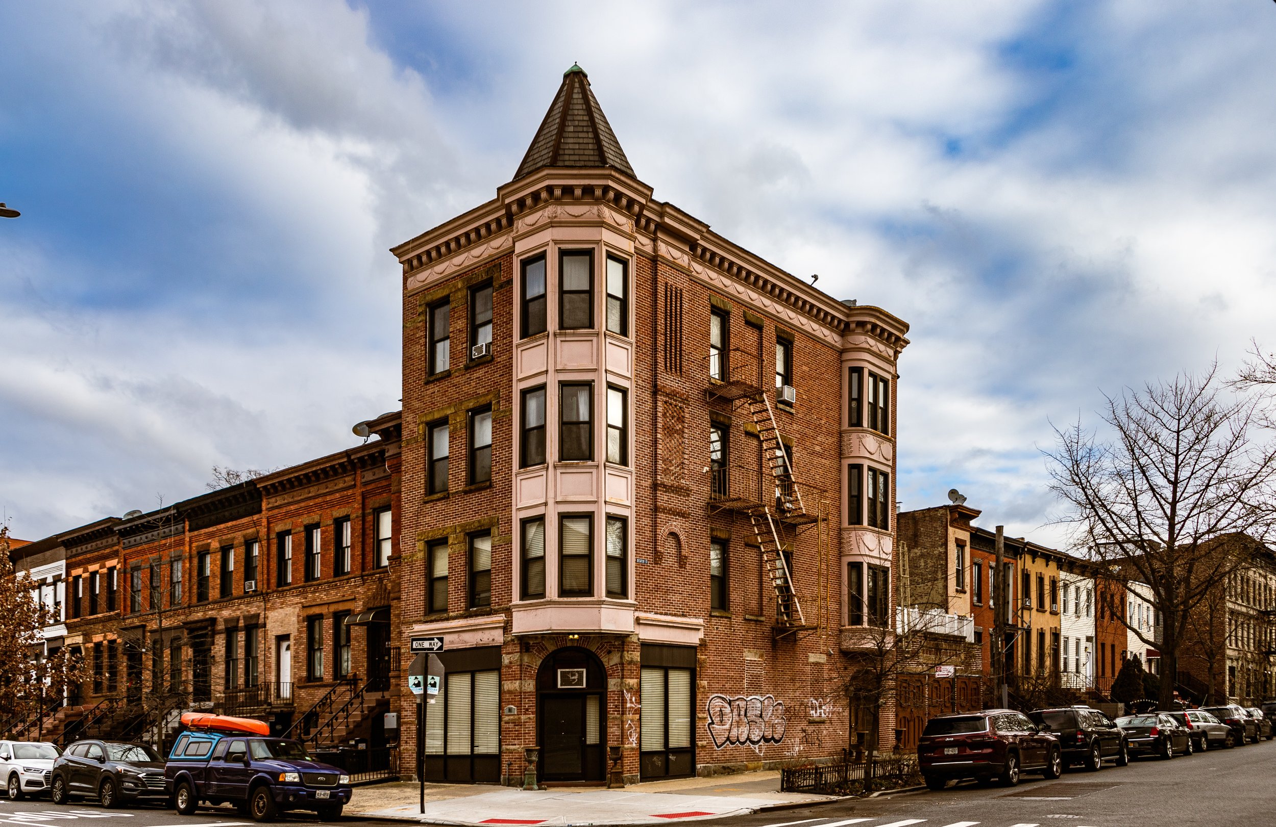 A brick apartment building with a turret on the corner, fire escapes on the facade, and a row of parked cars along the street, under a cloudy sky.