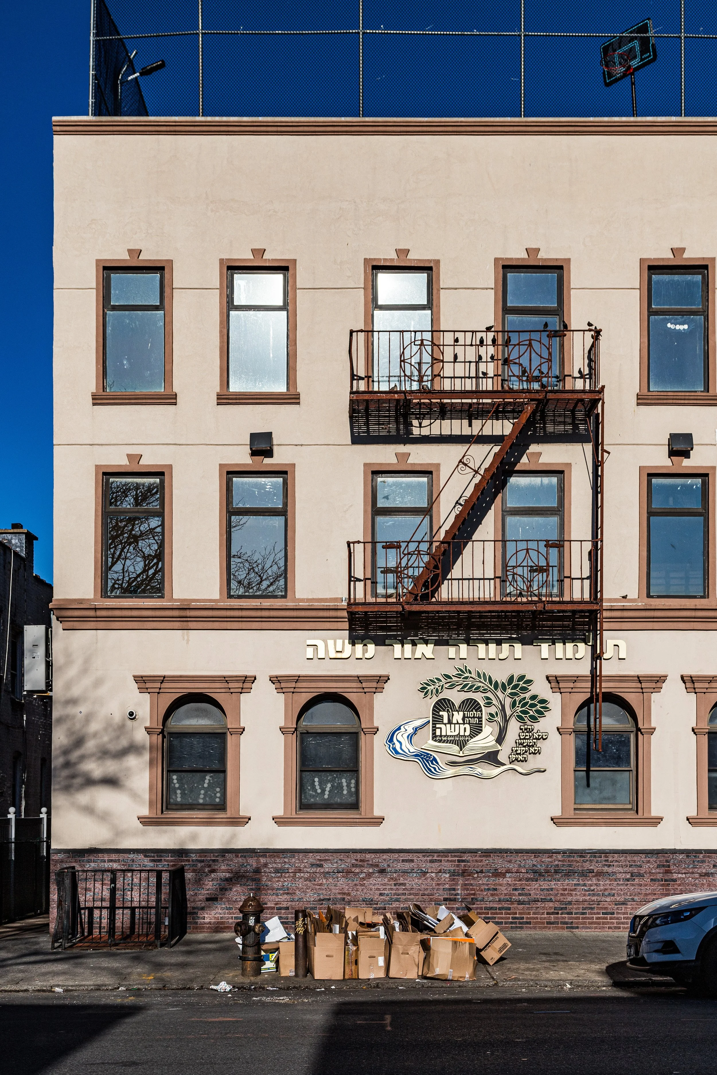 A four-story beige building with brown trim and a fire escape ladder on the front. The building has several windows and Hebrew writing on the wall along with a logo. There are boxes of items and a fire hydrant outside in front of the building.