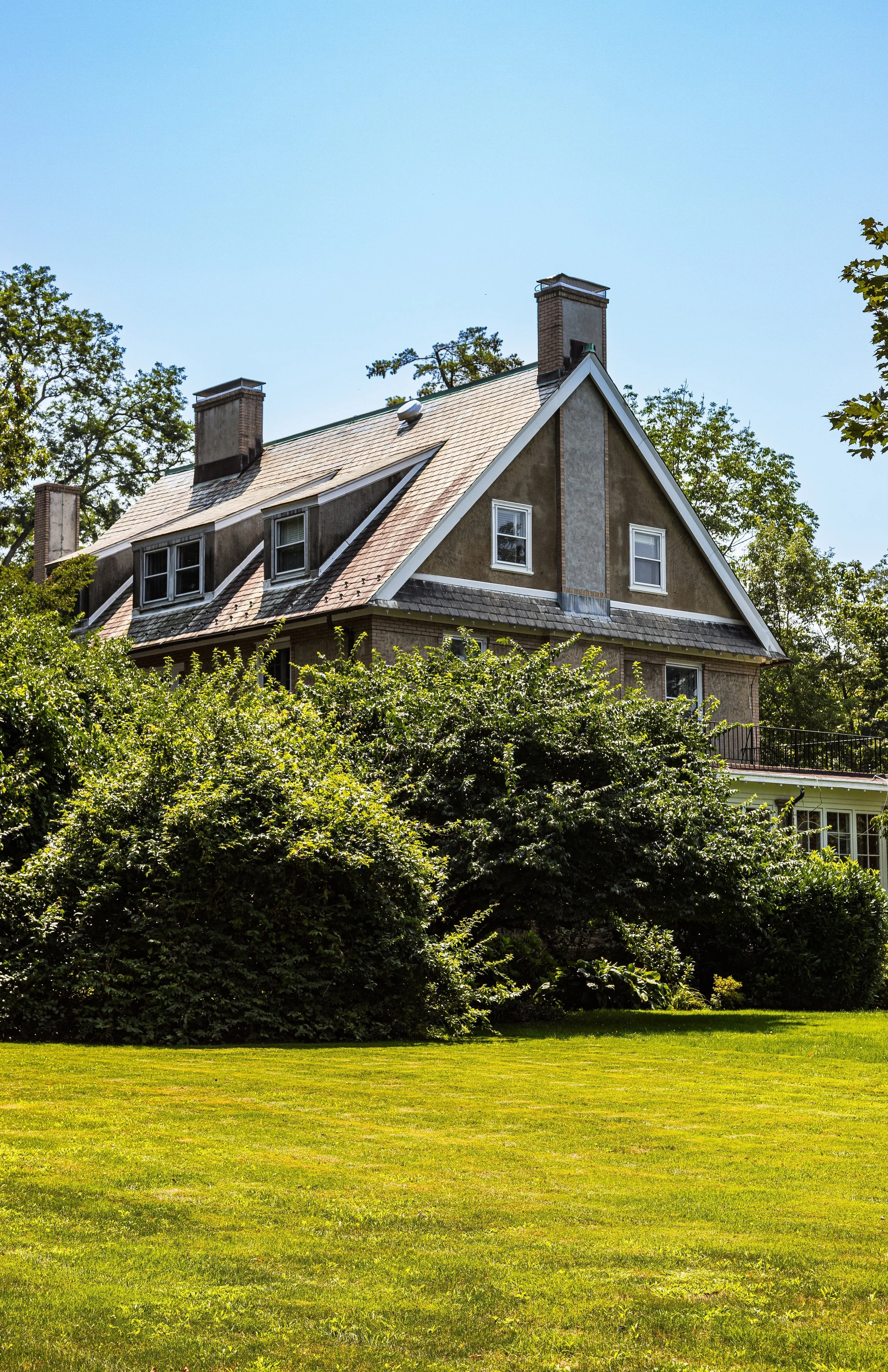 A house with a steep gabled roof, multiple chimneys, and several windows, surrounded by green trees and a well-maintained lawn under a clear blue sky.