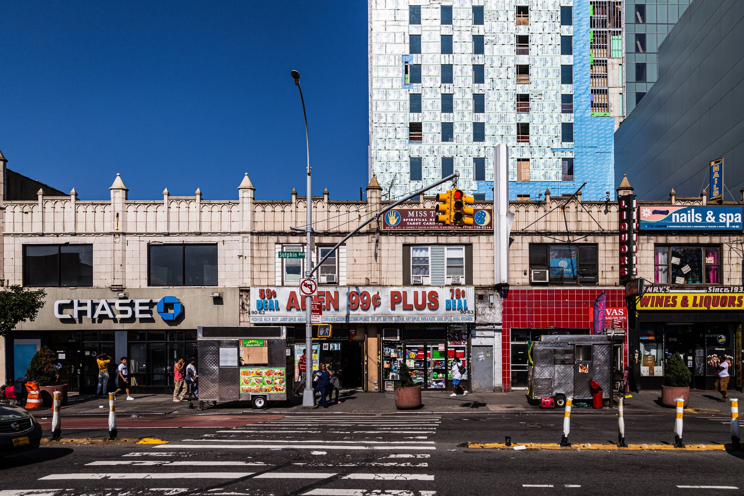 Urban street scene with multiple storefronts, including Chase bank, a dollar store, nail and spa salon, liquor store, and food vendors, under a clear blue sky.