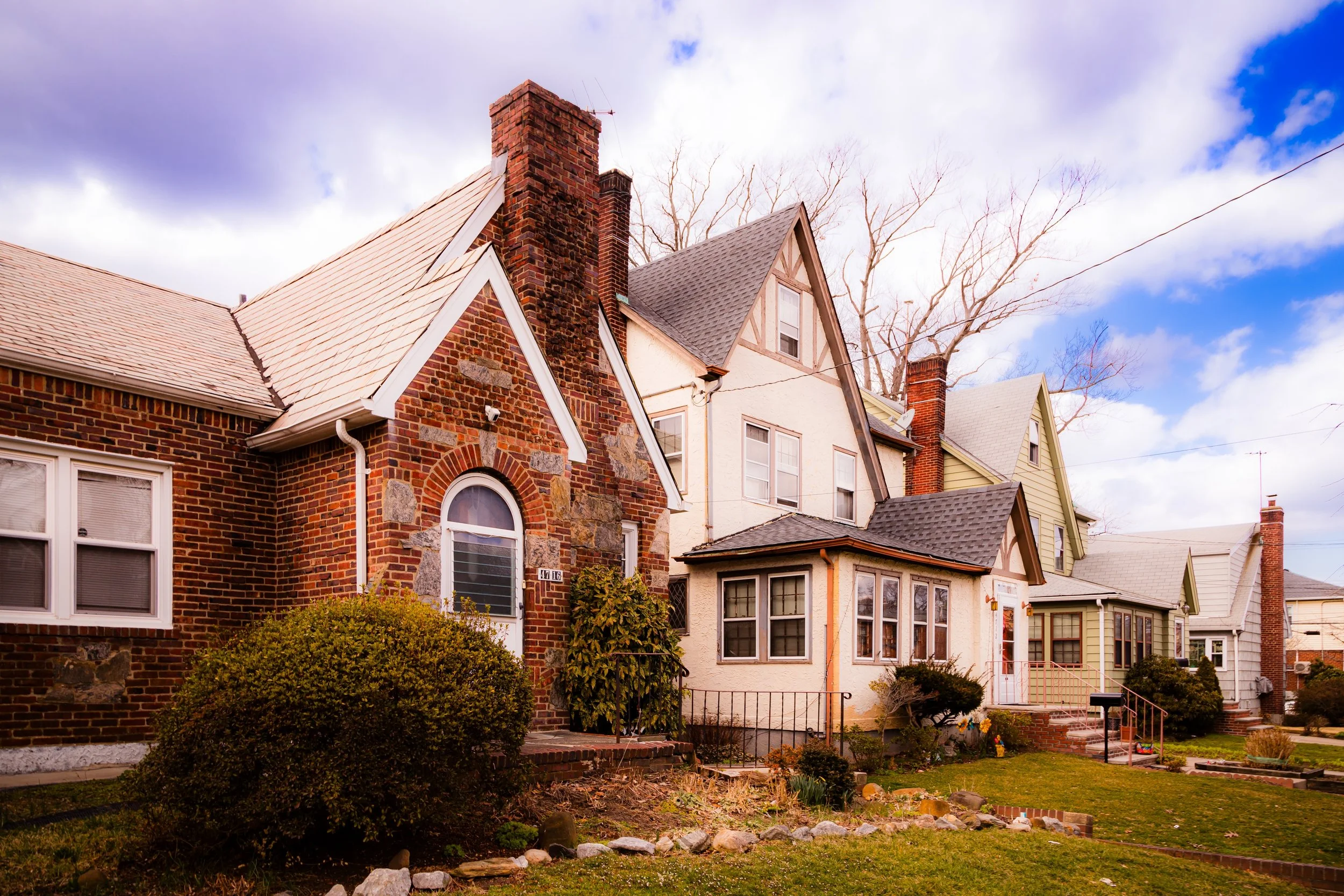 Row of historic brick and wood houses with gabled roofs and front yards under a partly cloudy sky.