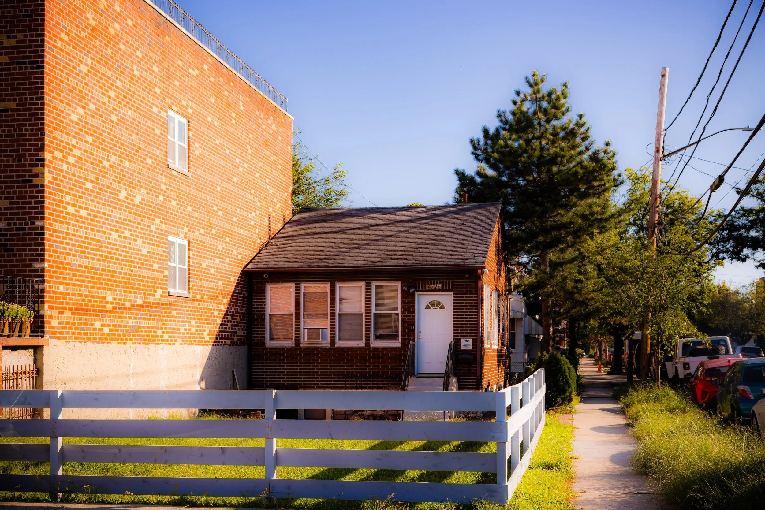 A small brick house with four windows and a white door, surrounded by a white fence, adjacent to a sidewalk with cars parked along it, and tall trees with green leaves, in a sunny neighborhood.