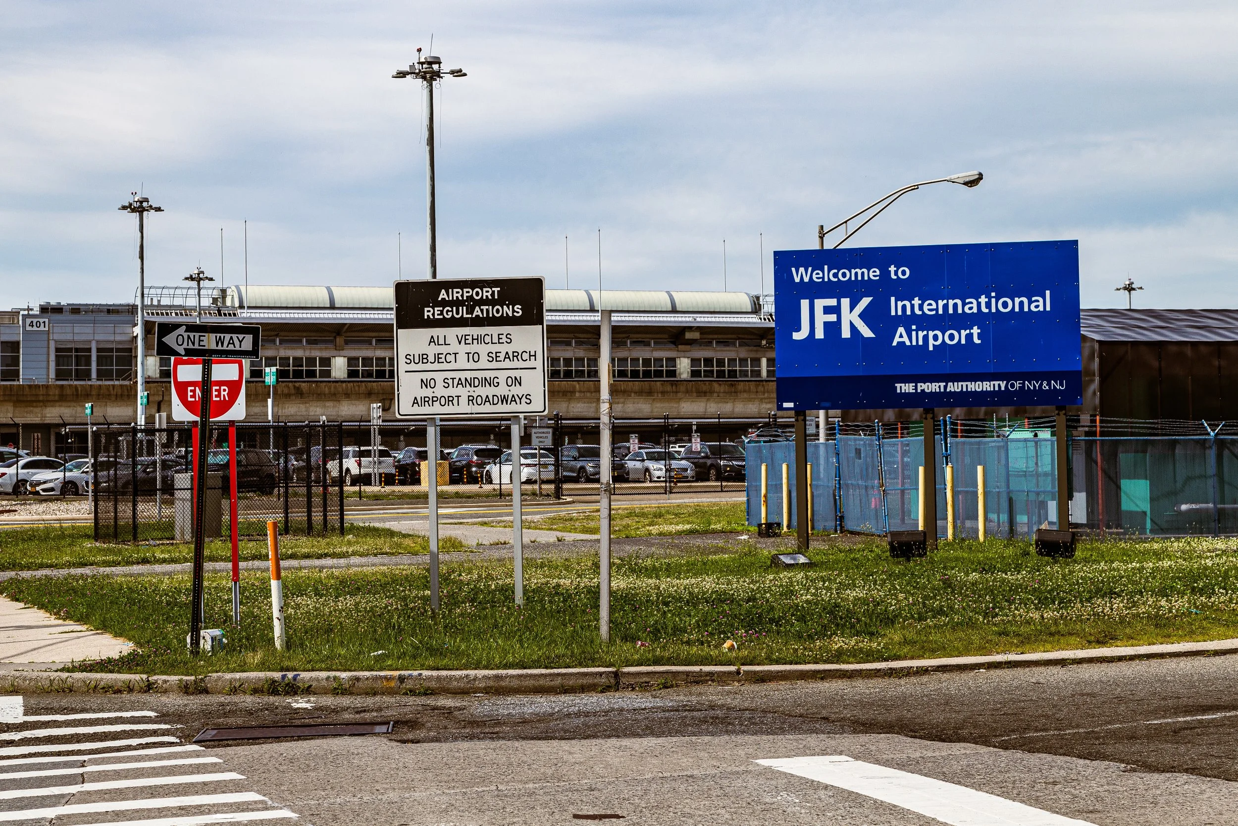 Sign at JFK International Airport entrance with parking lot, fence, and airport building in background.