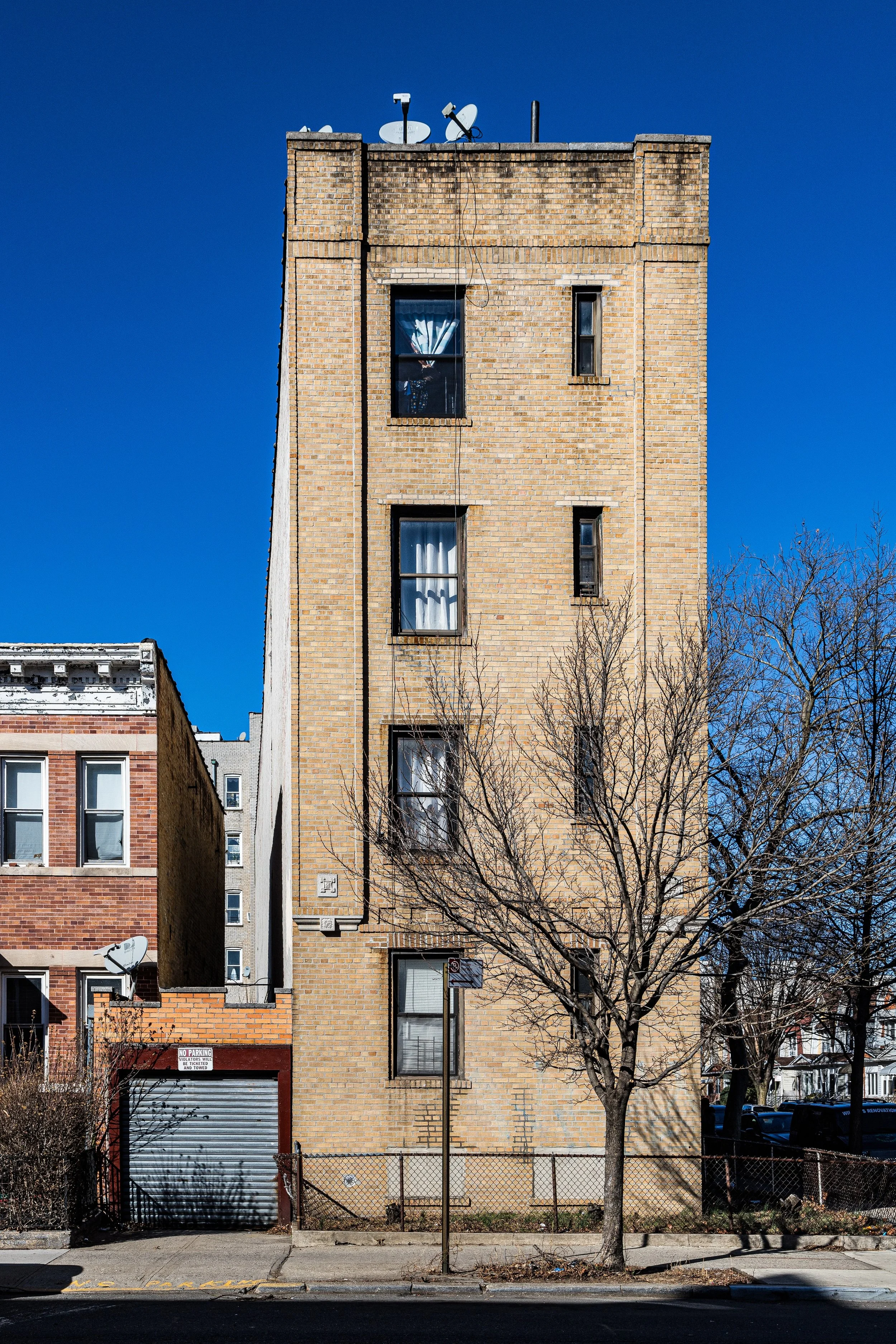 A multi-story brick building with four visible windows, some with curtains, and satellite dishes on the roof. A bare tree stands in front, with a sidewalk and a chain-link fence at the base.
