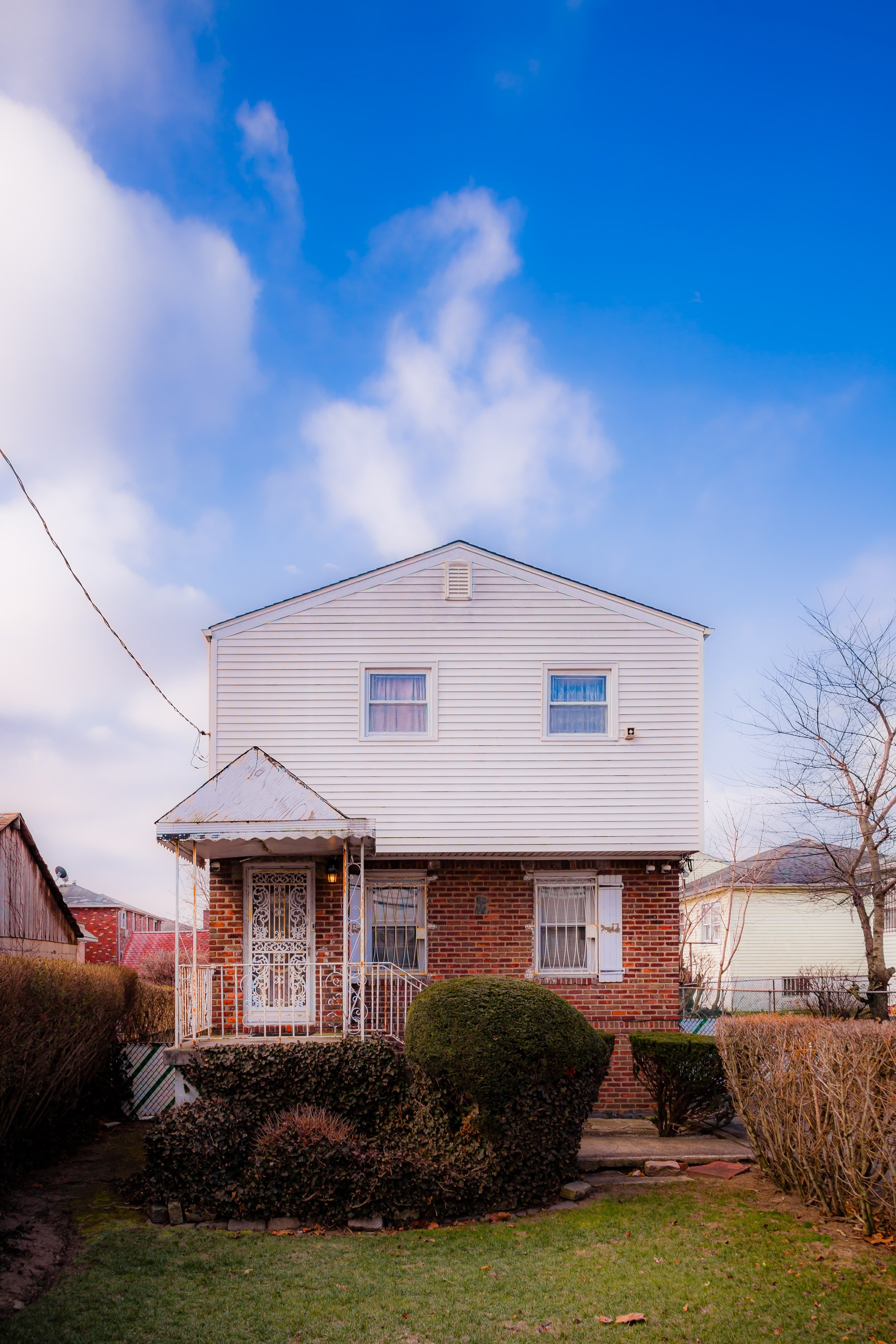 A two-story house with brick on the first floor and white siding on the second floor, with a small front porch, surrounded by a lawn and shrubs, under a partly cloudy blue sky.