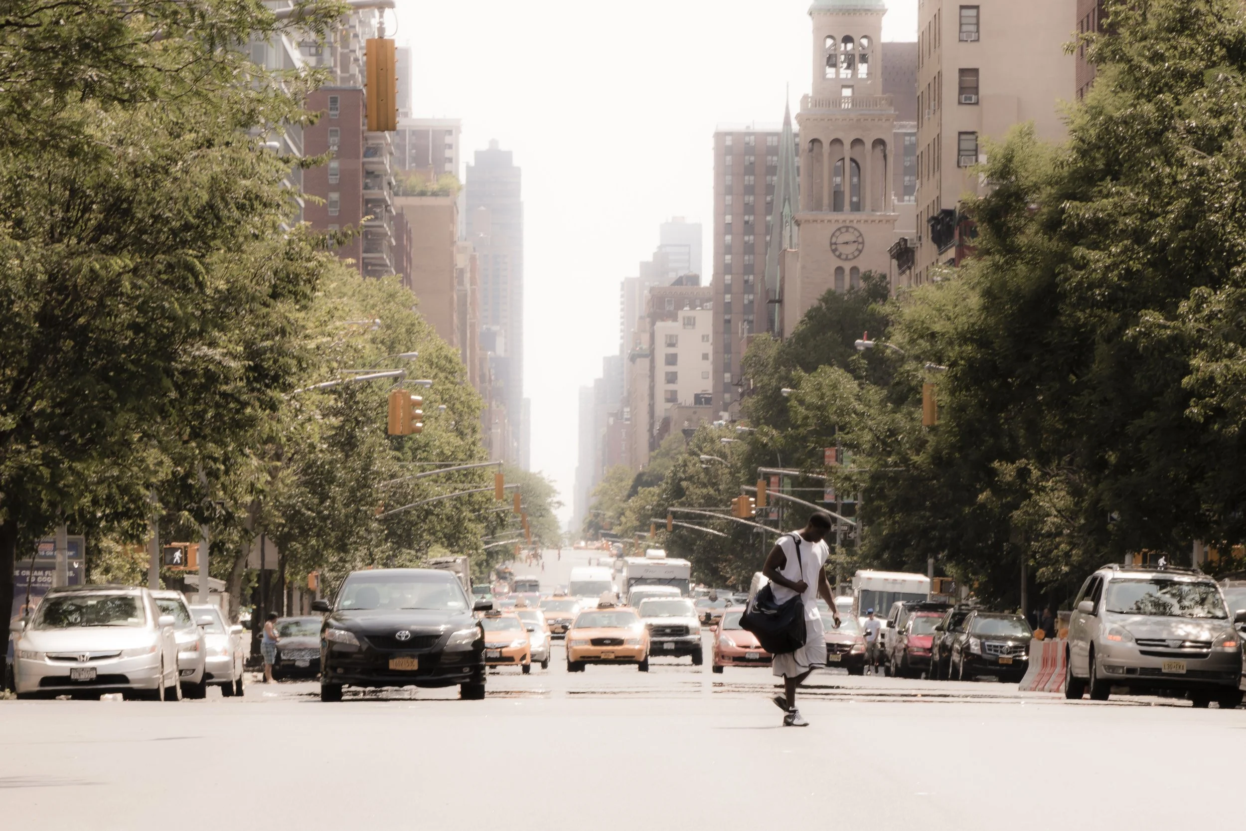 A person crossing a city street during daylight with cars parked and moving on both sides, tall buildings on the sides, trees lining the street, and a clock on a church tower in the background.