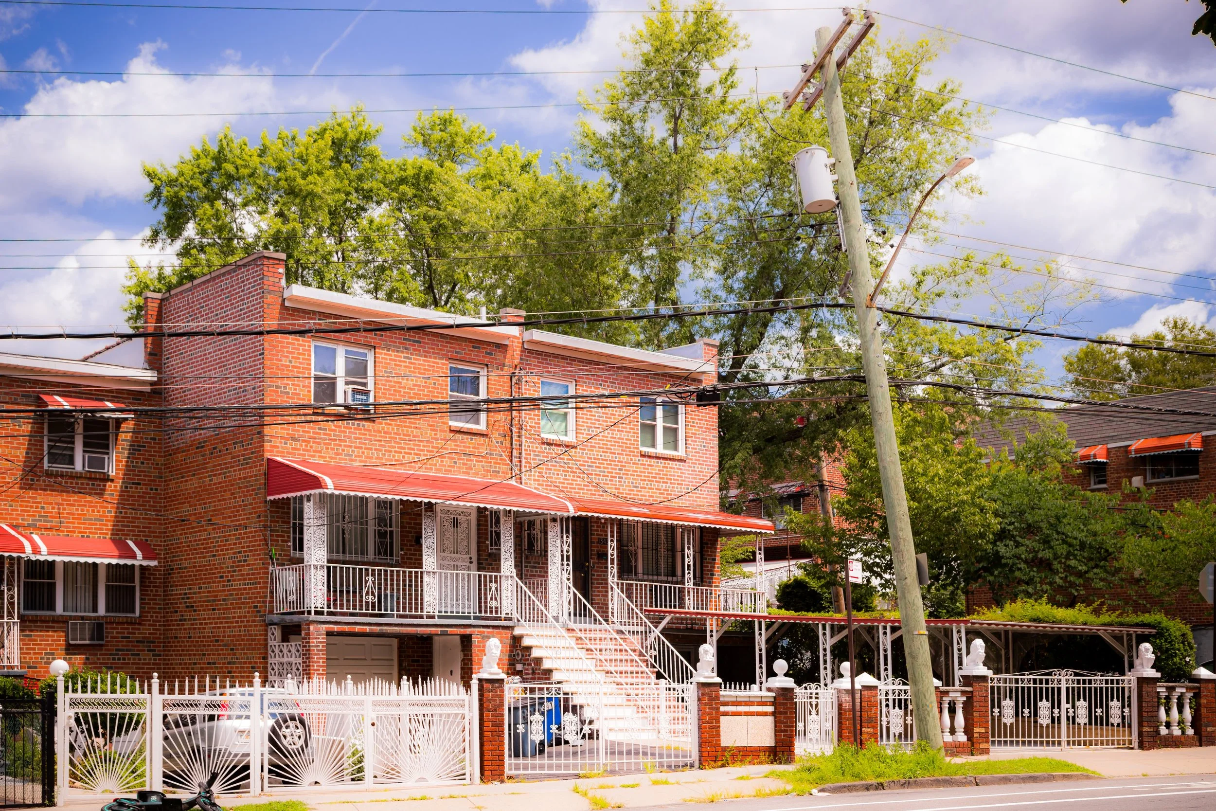 A two-story brick house with a white metal staircase leading to the front porch, decorated with white railings. The house has red roof accents and a white front door. A white fence surrounds the property, and there are trees and a utility pole in the front. The sky is partly cloudy with patches of blue.