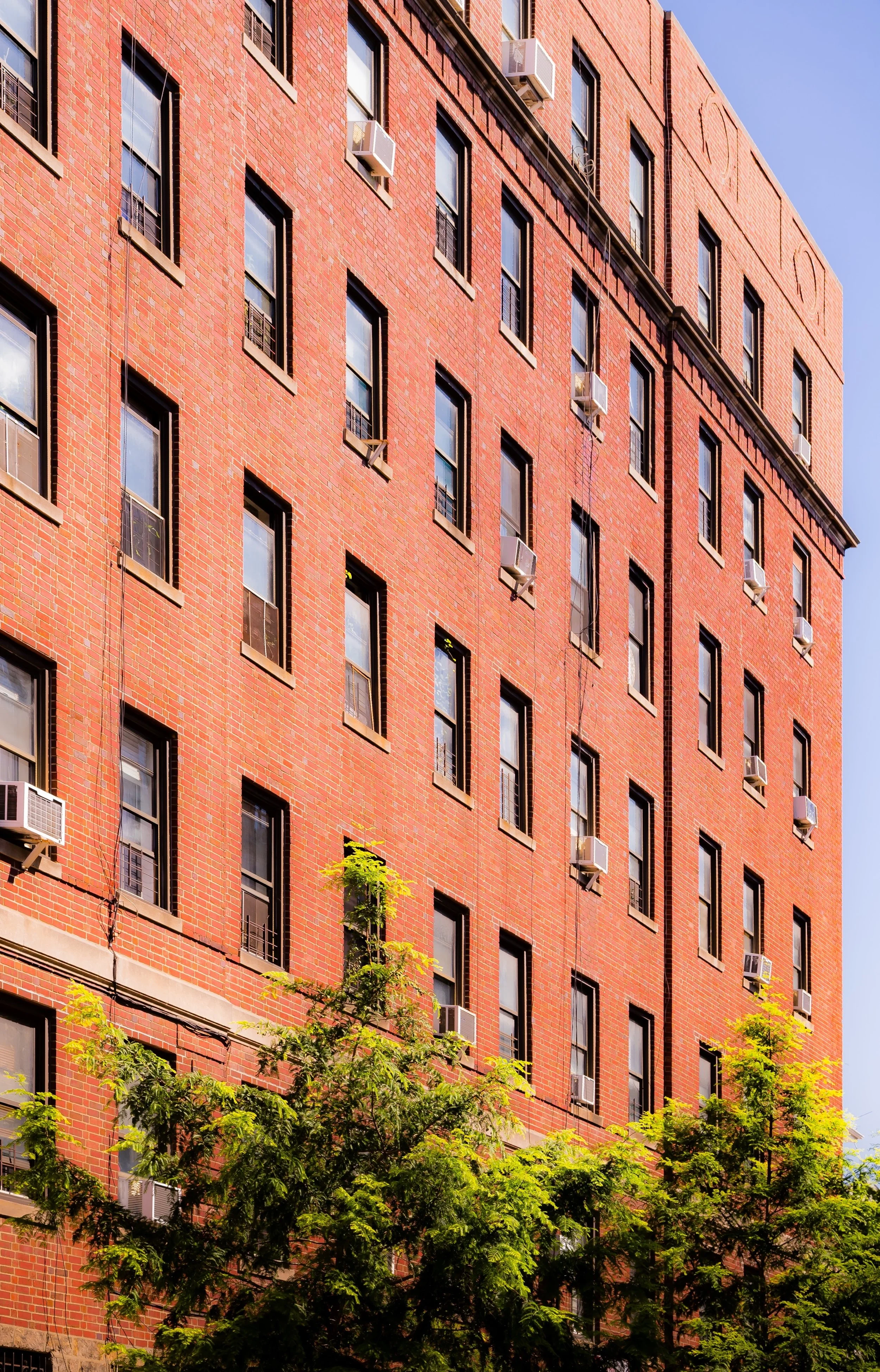 Red brick apartment building with multiple windows and air conditioning units, with green trees at the bottom and a clear blue sky above.