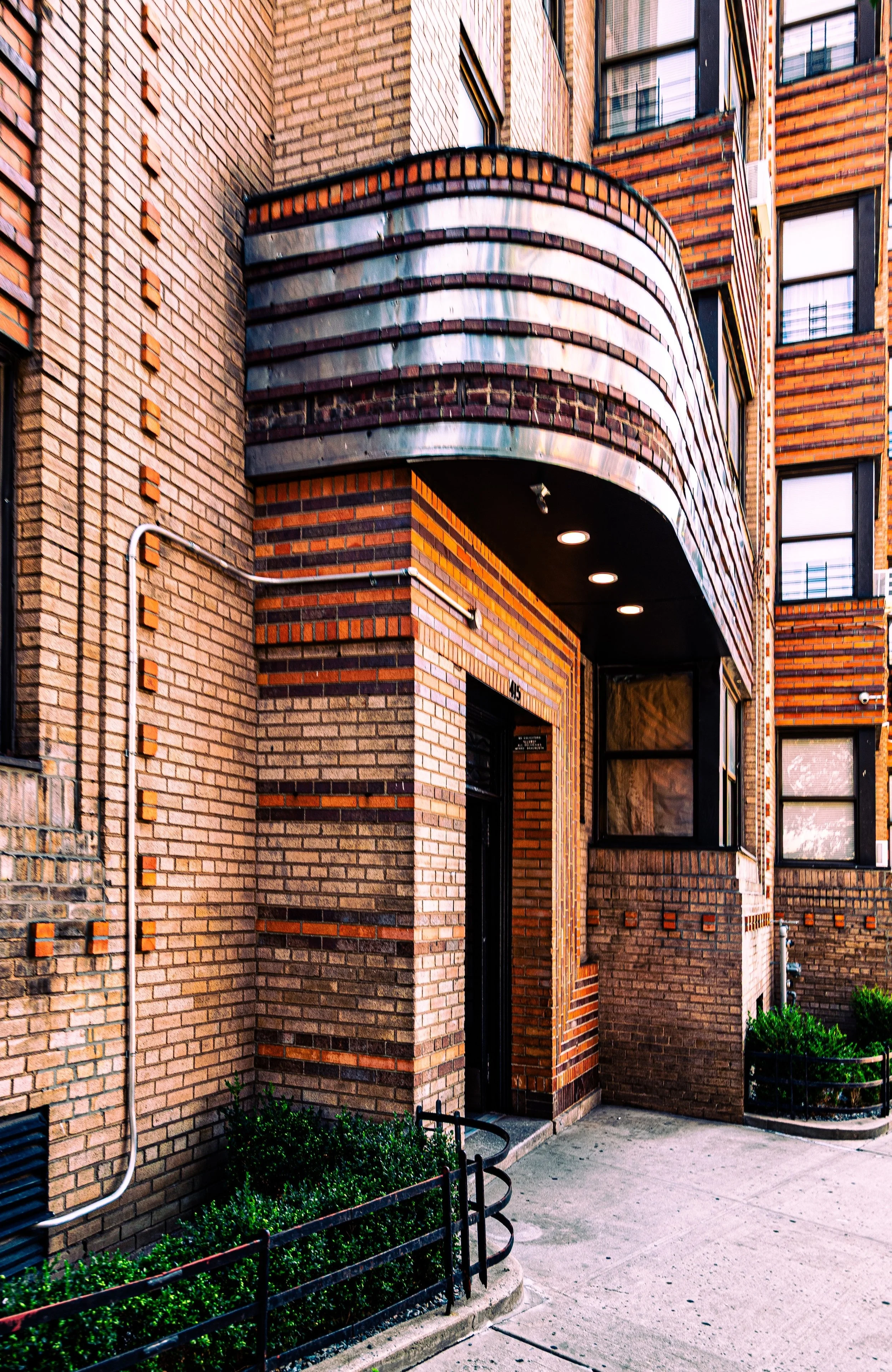 Facade of a modern brick apartment building with an overhang covered in metallic strips, illuminated by a row of recessed ceiling lights, with a sidewalk and small garden patch at the entrance.