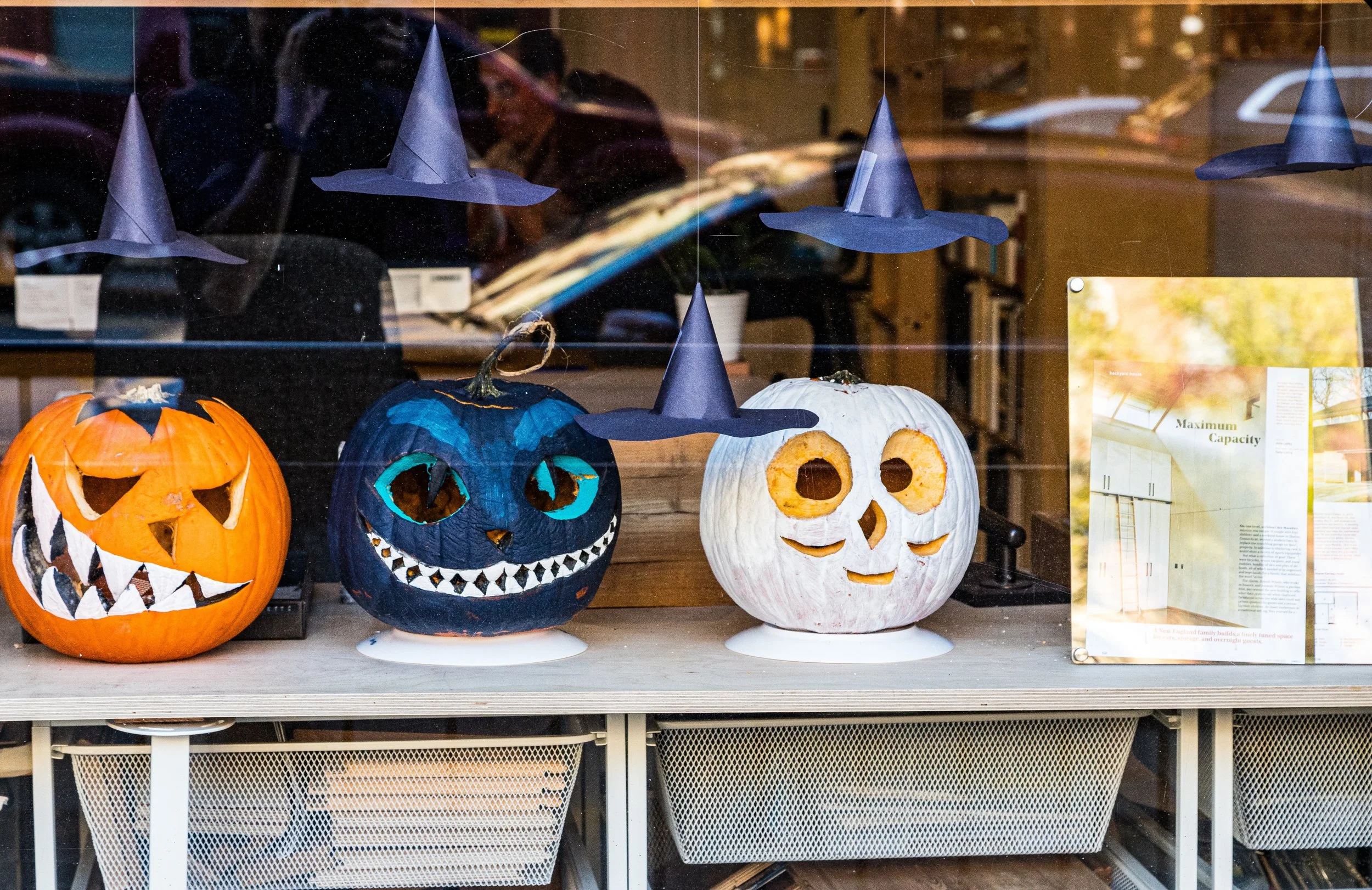 Display of three Halloween-themed pumpkins with carved faces decorated with witch hats, set inside a shop window with reflections of the street and a sign/board with text behind the glass.