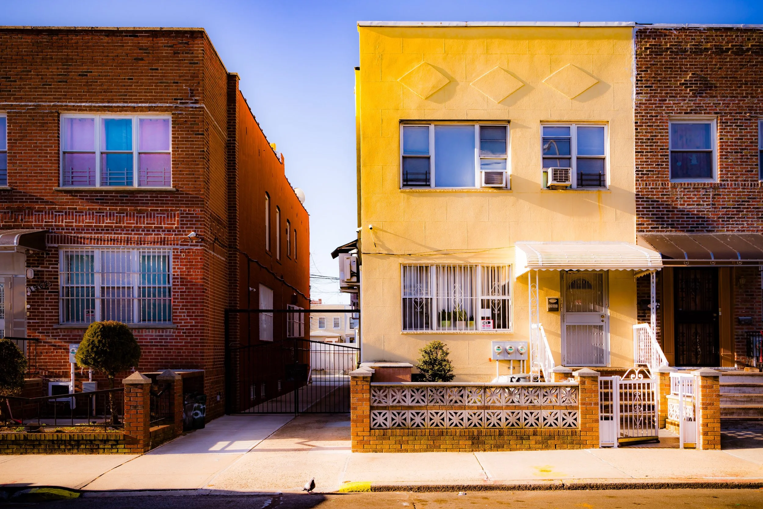 Three townhouse-style buildings on a city street, with brick and yellow walls, windows with bars and air conditioning units, small front yards, and a gate at the alleyway between them.