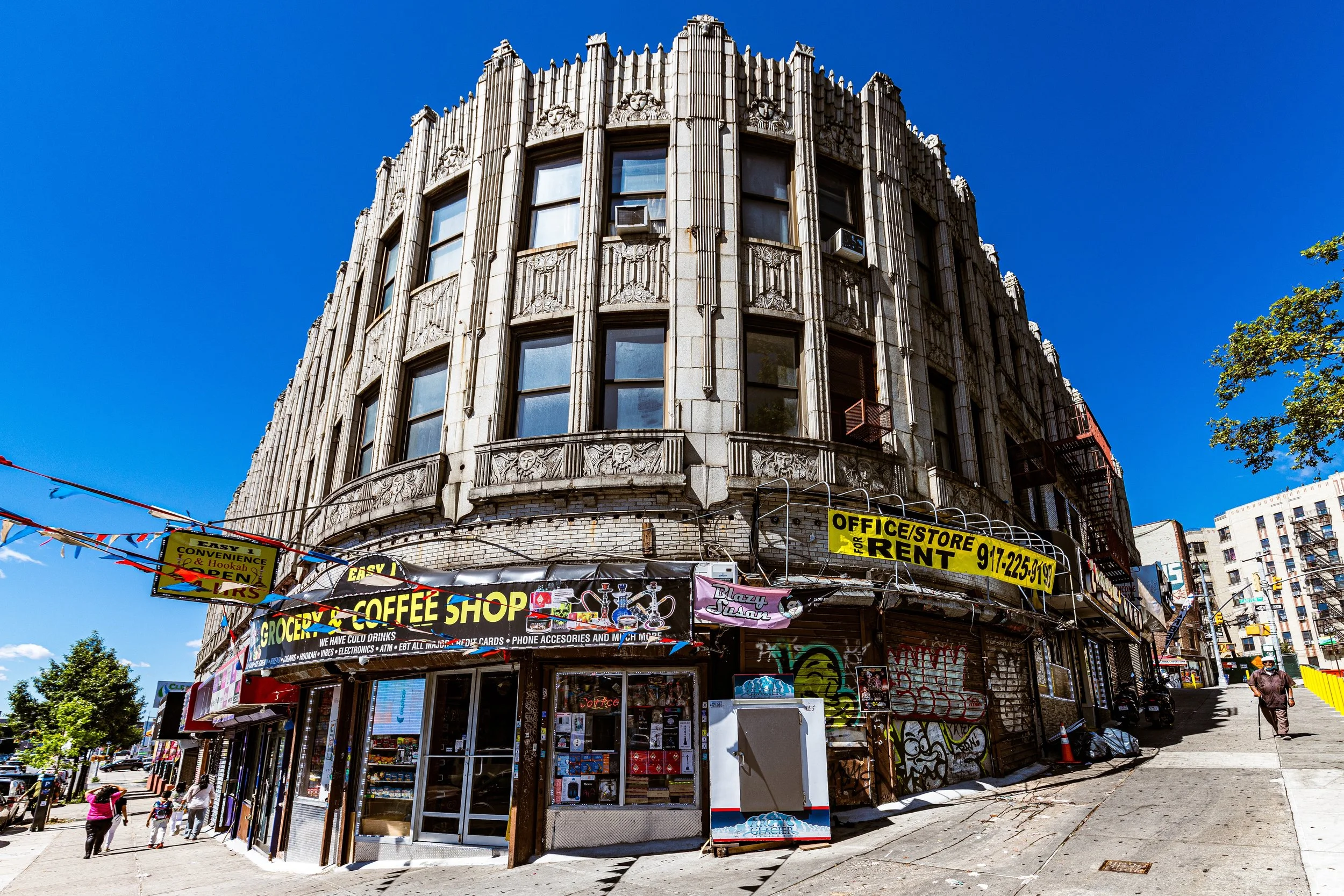 A vintage building with detailed architectural designs on top, housing a grocery store, coffee shop, convenience store, and offices for rent, with colorful street signs and graffiti, on a street with pedestrians, trees, and another building in the background under a clear blue sky.