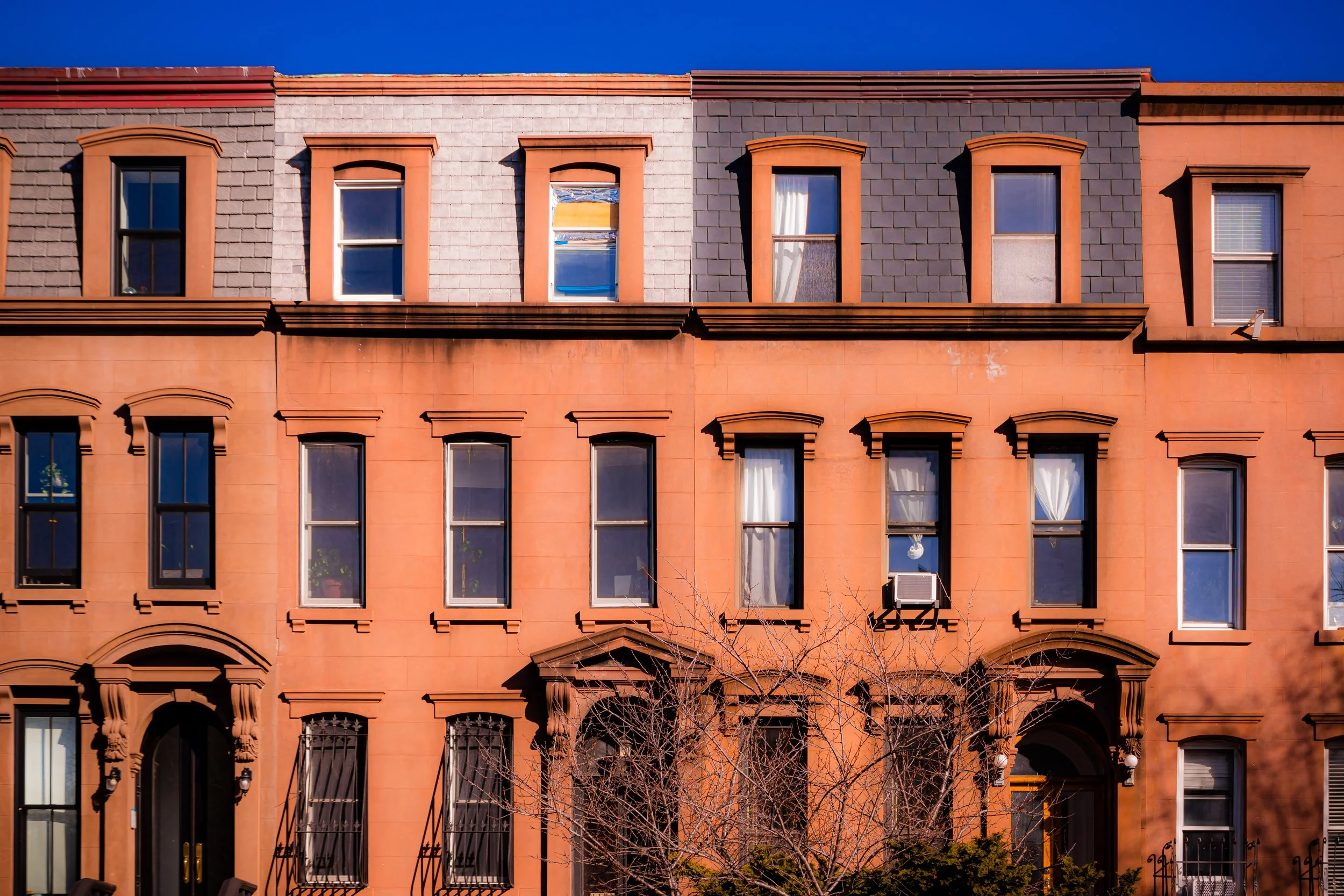 Brownstone row house with multiple tall windows and decorative arches, some with curtains or air conditioning units, leafless trees in front, under a clear blue sky.