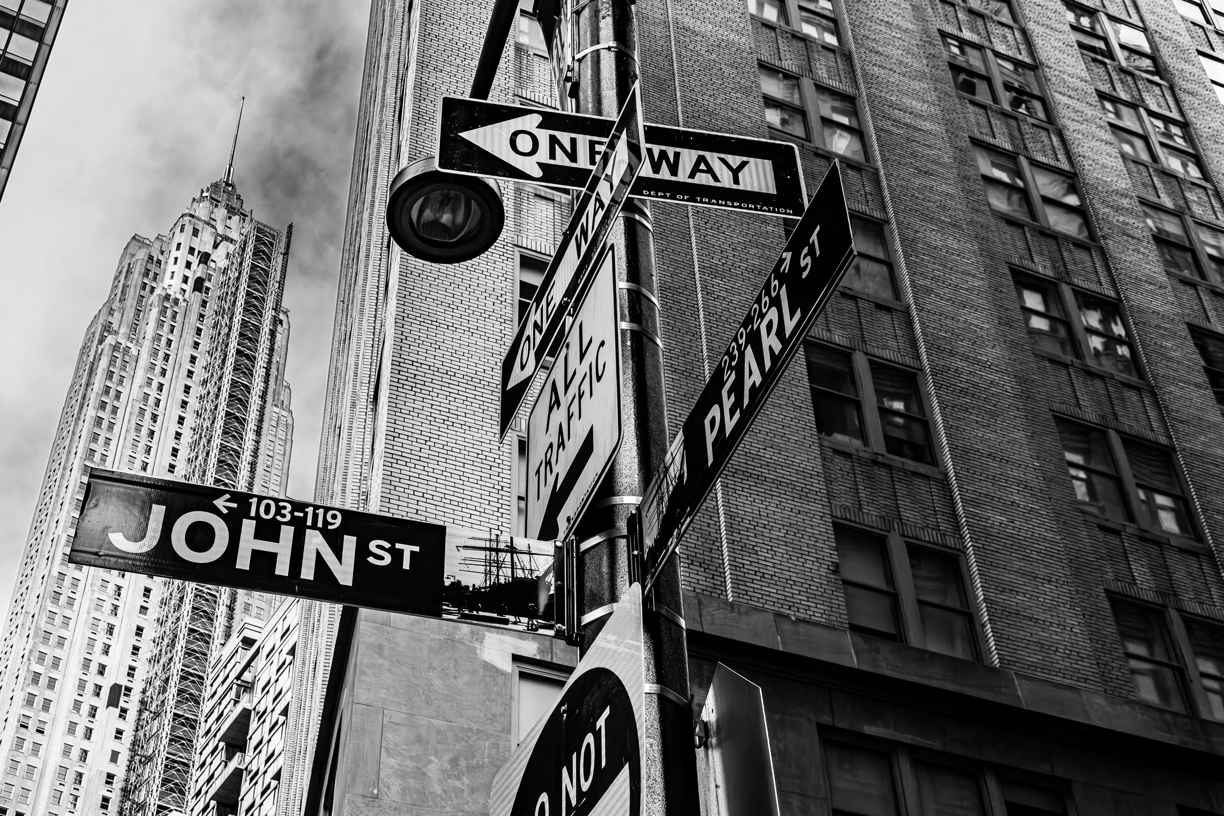 Street signs at an intersection in New York City, including a one way sign, a street name sign for John Street, and signs indicating traffic directions and parking restrictions, with tall buildings in the background.