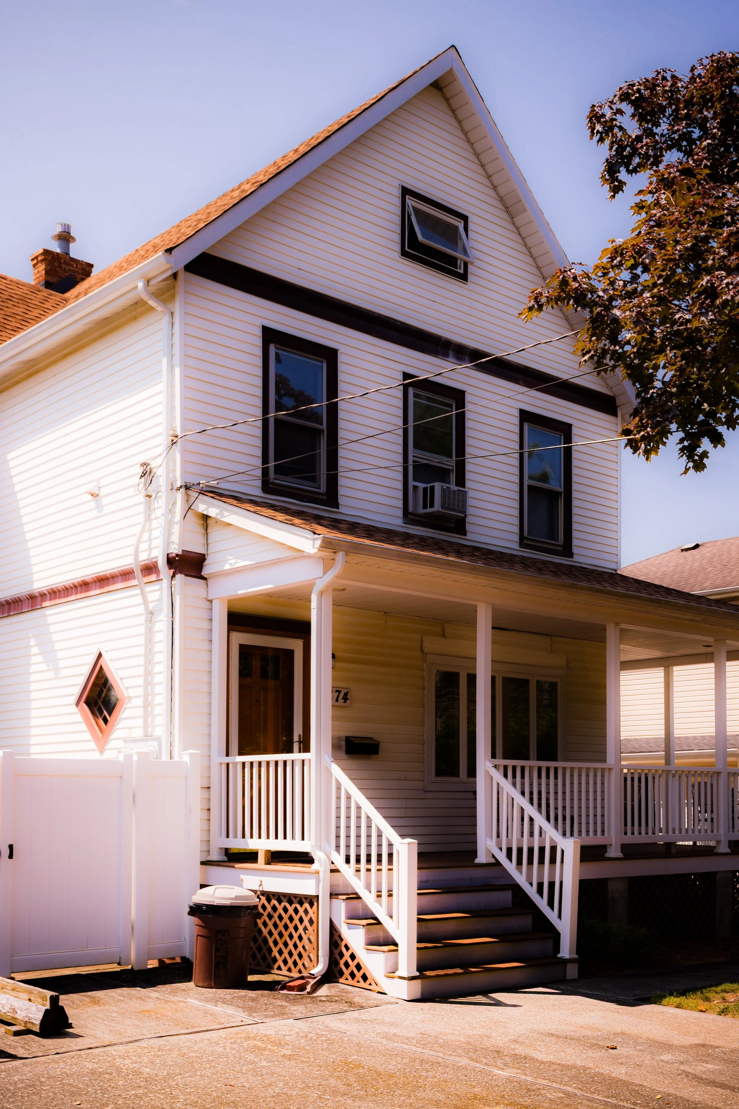 A two-story white house with a front porch, stairs, and an exterior air conditioning unit near a window, with a tree on the right side and a clear sky overhead.