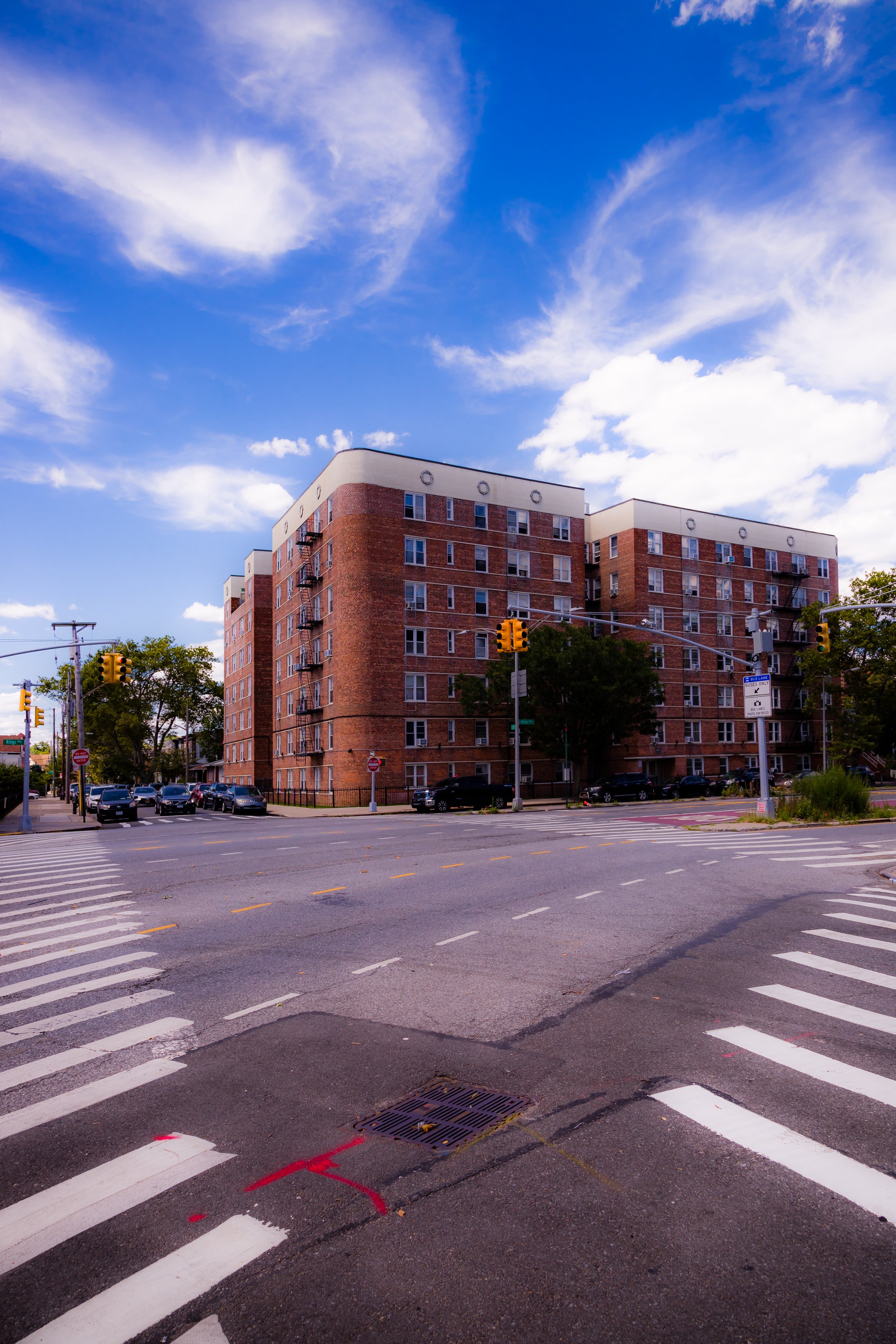 A city intersection with a multi-story brick apartment building in the background, traffic lights, cars parked along the street, and a pedestrian crosswalk under a partly cloudy blue sky.