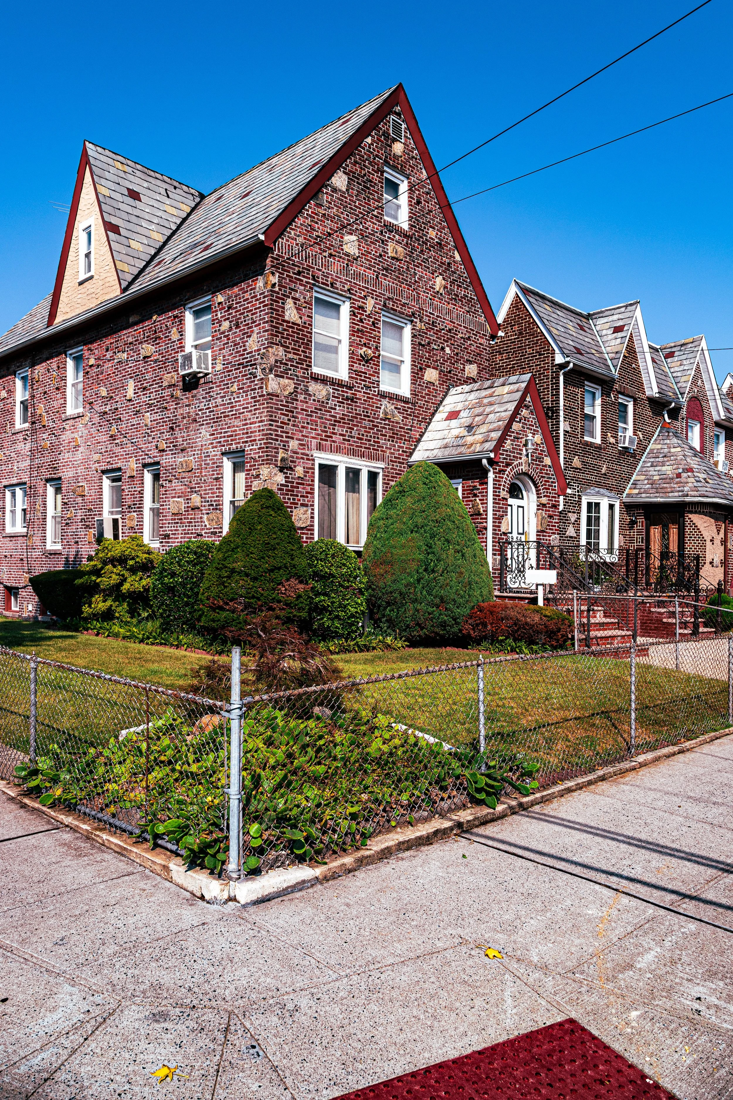 A multi-story brick residential building with a pointed roof, small front yard with manicured bushes, and a sidewalk enclosed by a metal fence.