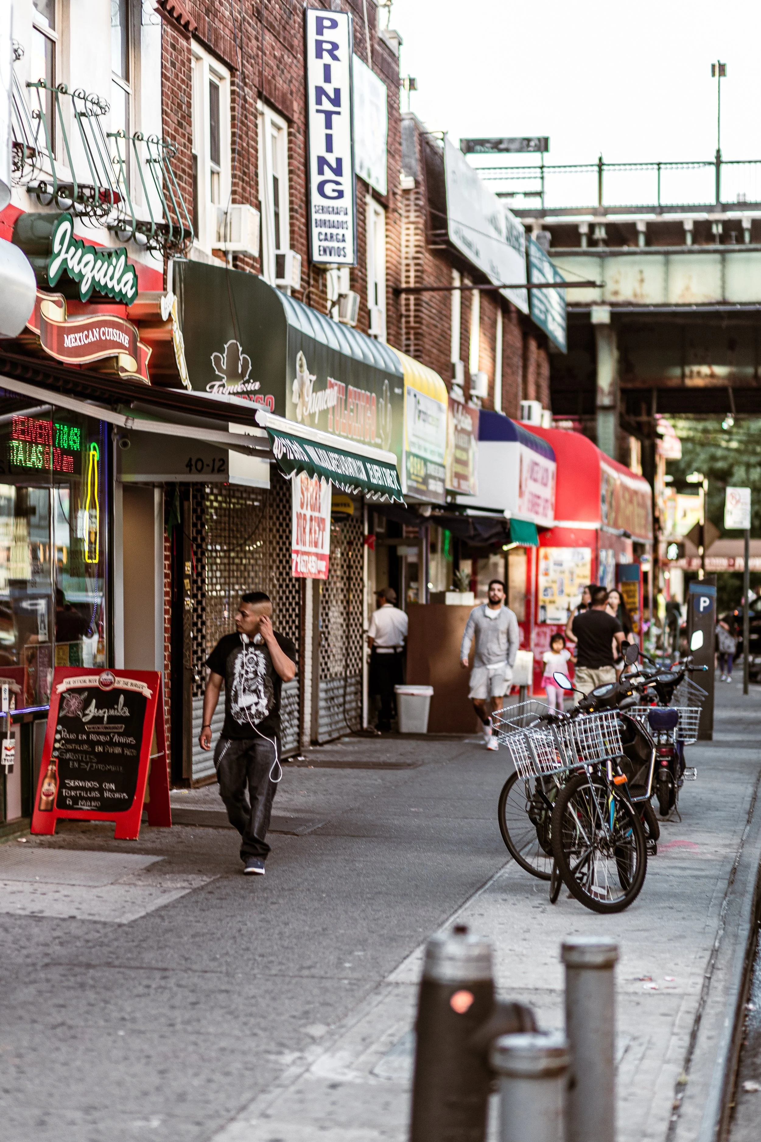 City street scene showing storefronts, bicycles, pedestrians, and signage for businesses including a printing shop, a Mexican restaurant, and others under a bridge or elevated railroad track.