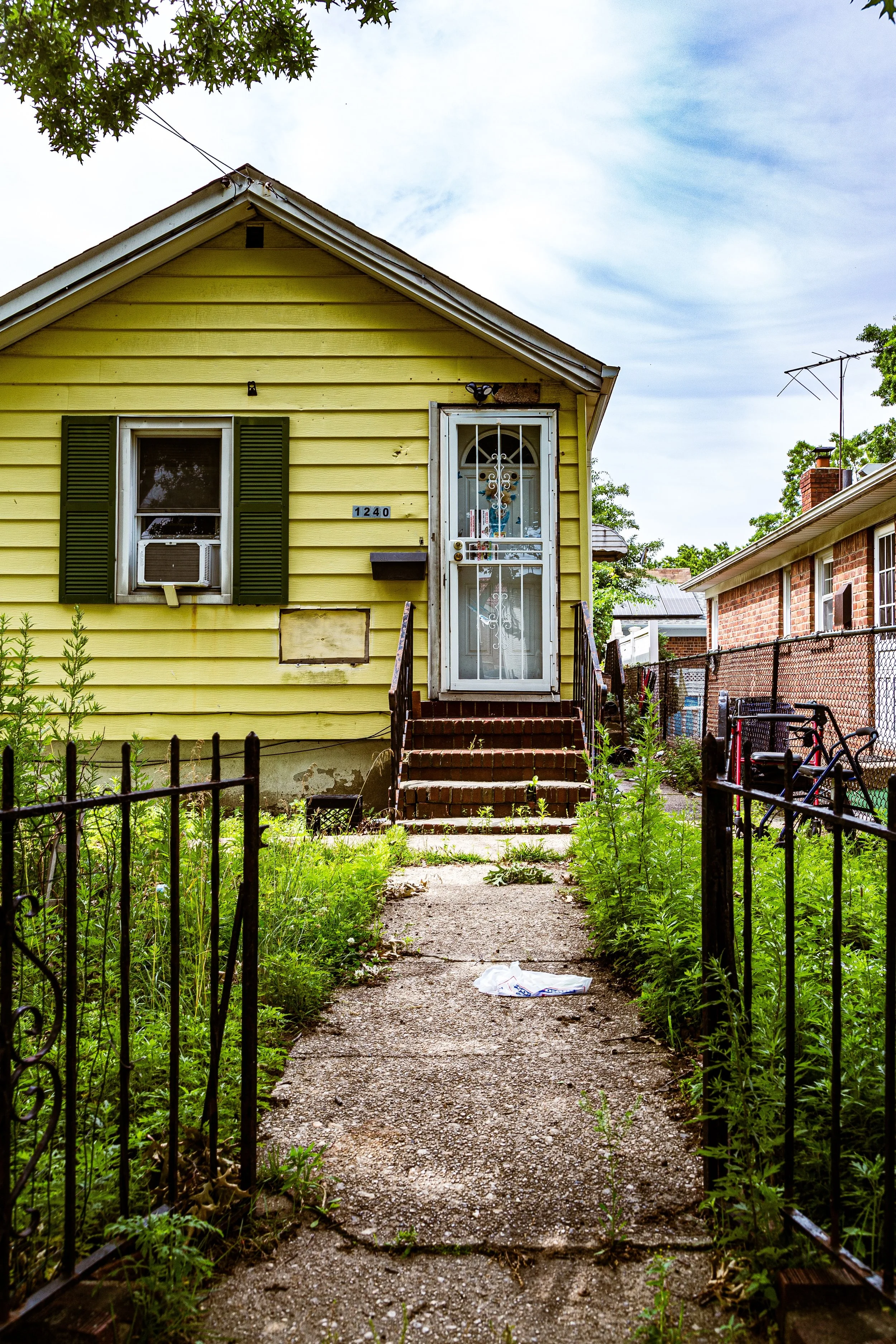 A yellow house with green shutters and a white security door, with a small front yard overgrown with weeds, a narrow cracked concrete sidewalk, and a black metal fence surrounding the yard, under a partly cloudy sky.
