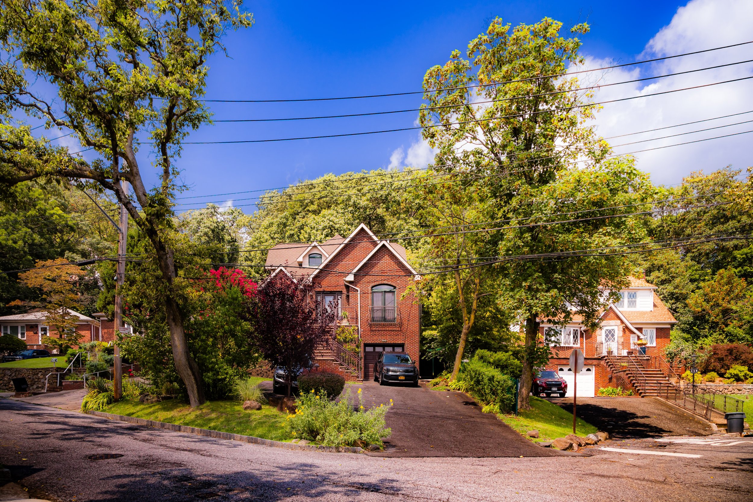 A suburban street with two houses surrounded by trees, parked cars, and blue skies.
