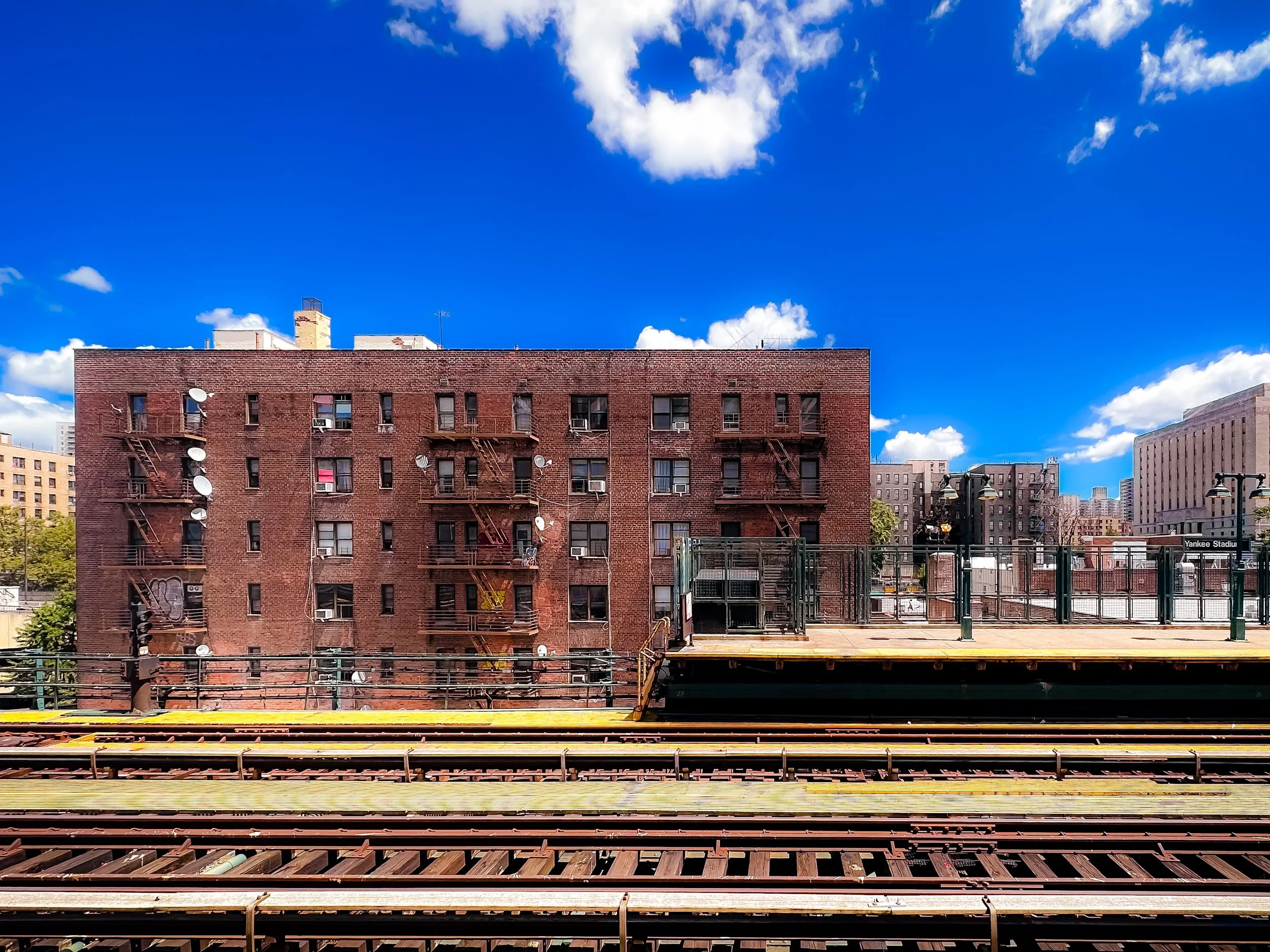 View of a red brick apartment building with fire escape staircases and satellite dishes, set against a bright blue sky with scattered white clouds, with train tracks in the foreground and other city buildings in the background.