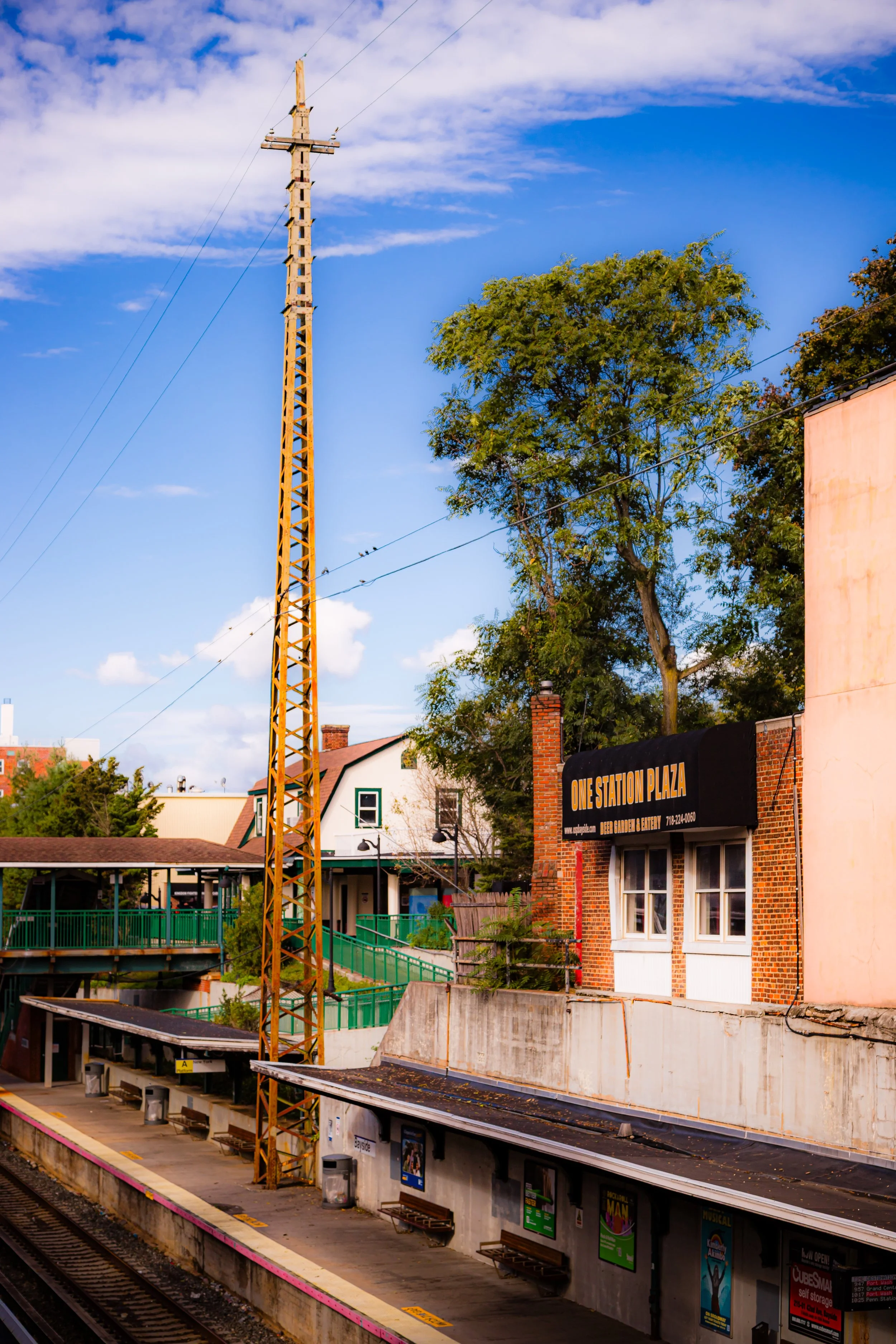 A train station platform with benches and posters, a tall yellow utility pole, trees, buildings, and a sign that reads 'One Station Plaza' under a partly cloudy sky.