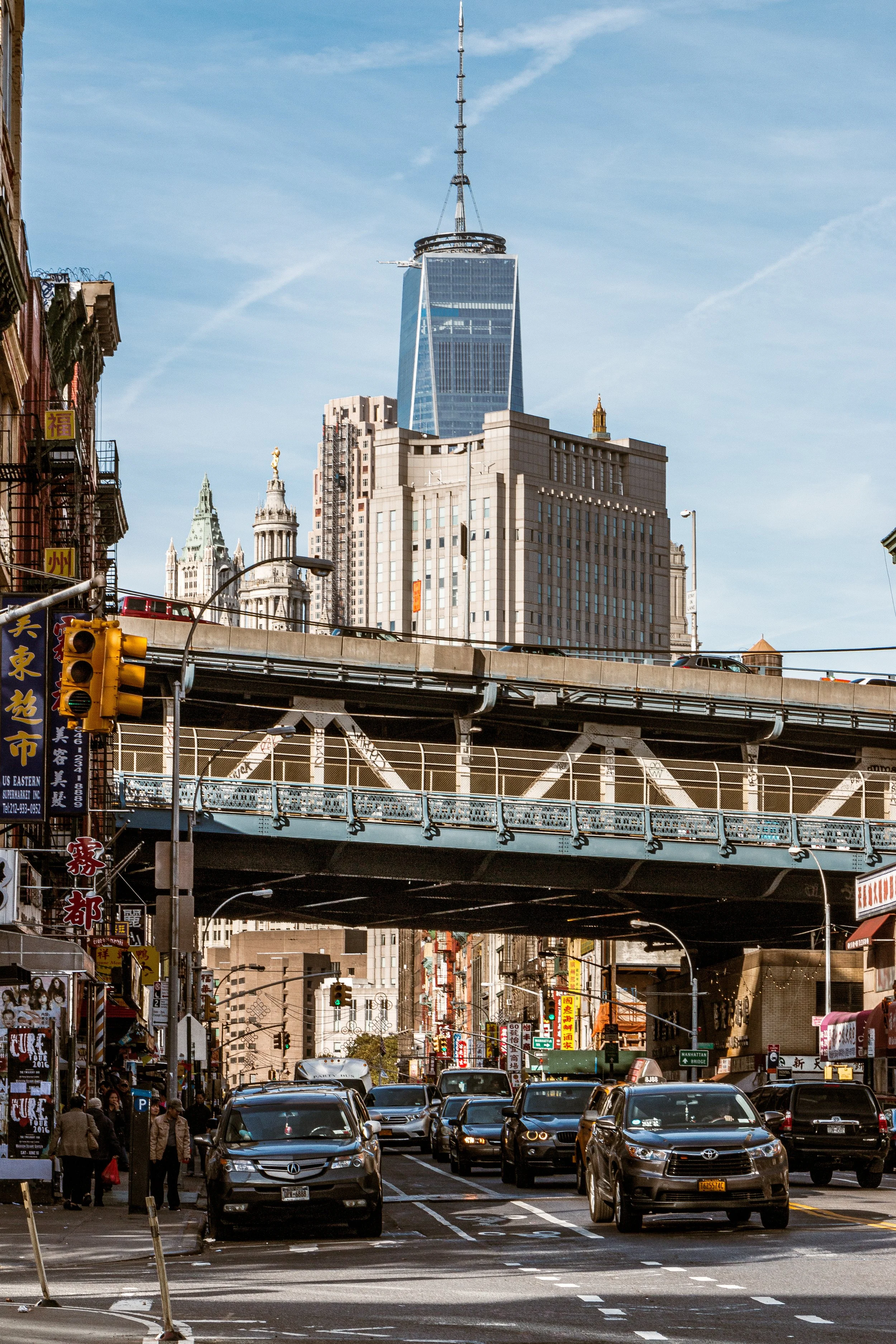 City street scene with cars stopped at a traffic light, Chinatown storefronts with signs in Chinese characters, and a background of high-rise buildings including One World Trade Center in New York City.