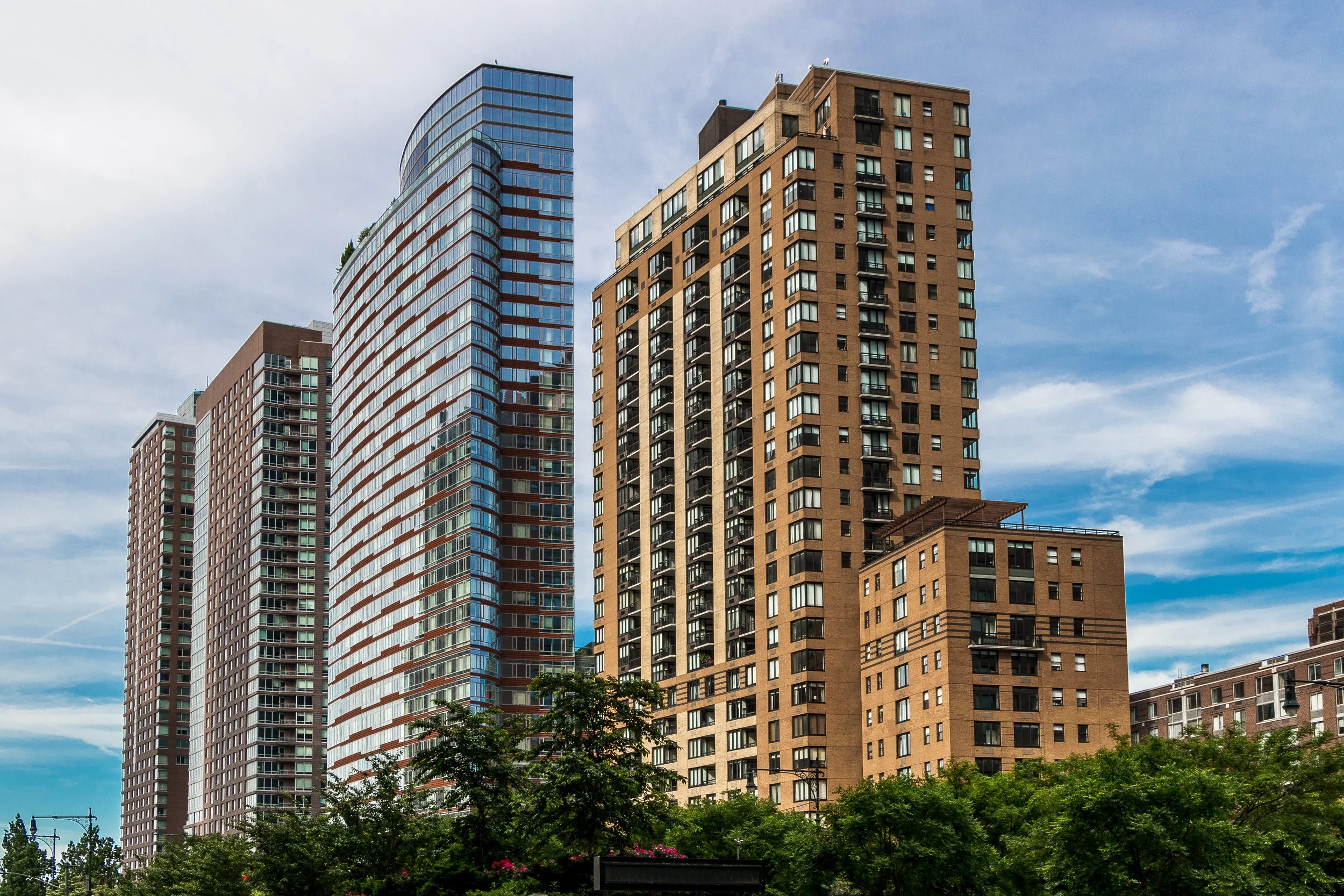 Tall modern high-rise buildings with glass and brick facades against a partly cloudy sky, with green trees at the base.