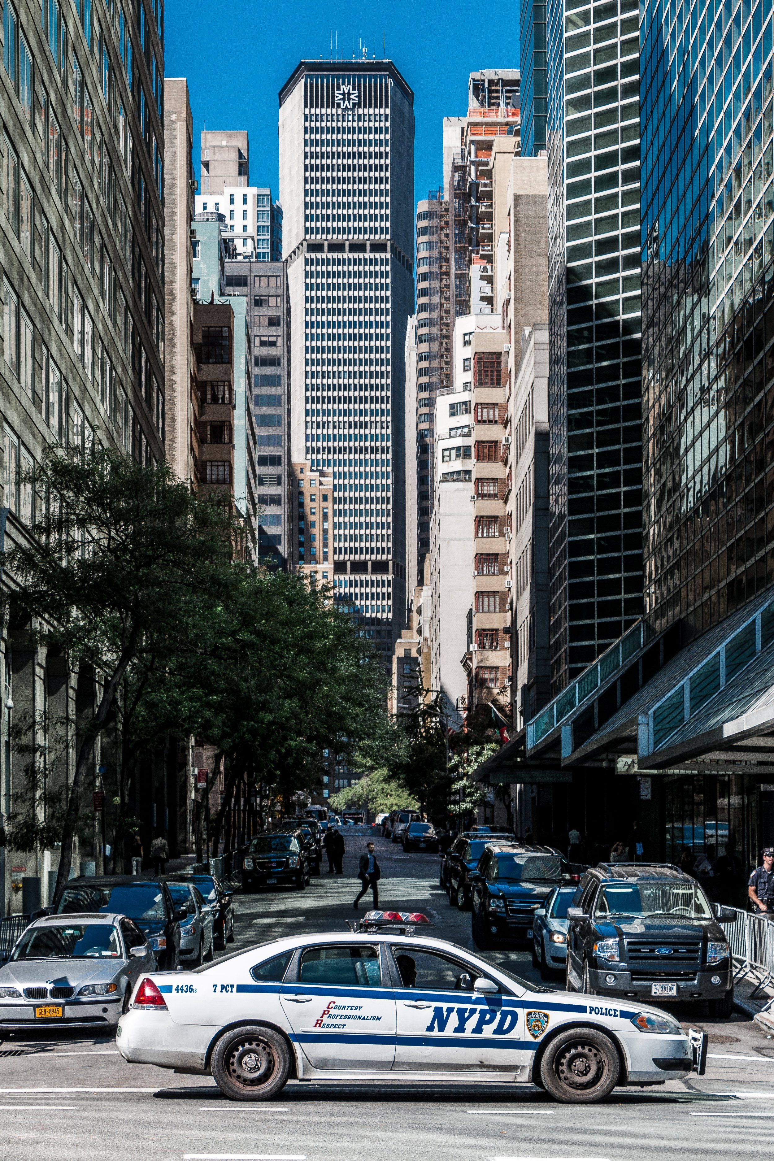 A city street scene with a white NYPD police car parked in the foreground, several cars parked along the street, pedestrians walking, and tall skyscrapers in clear blue sky background.