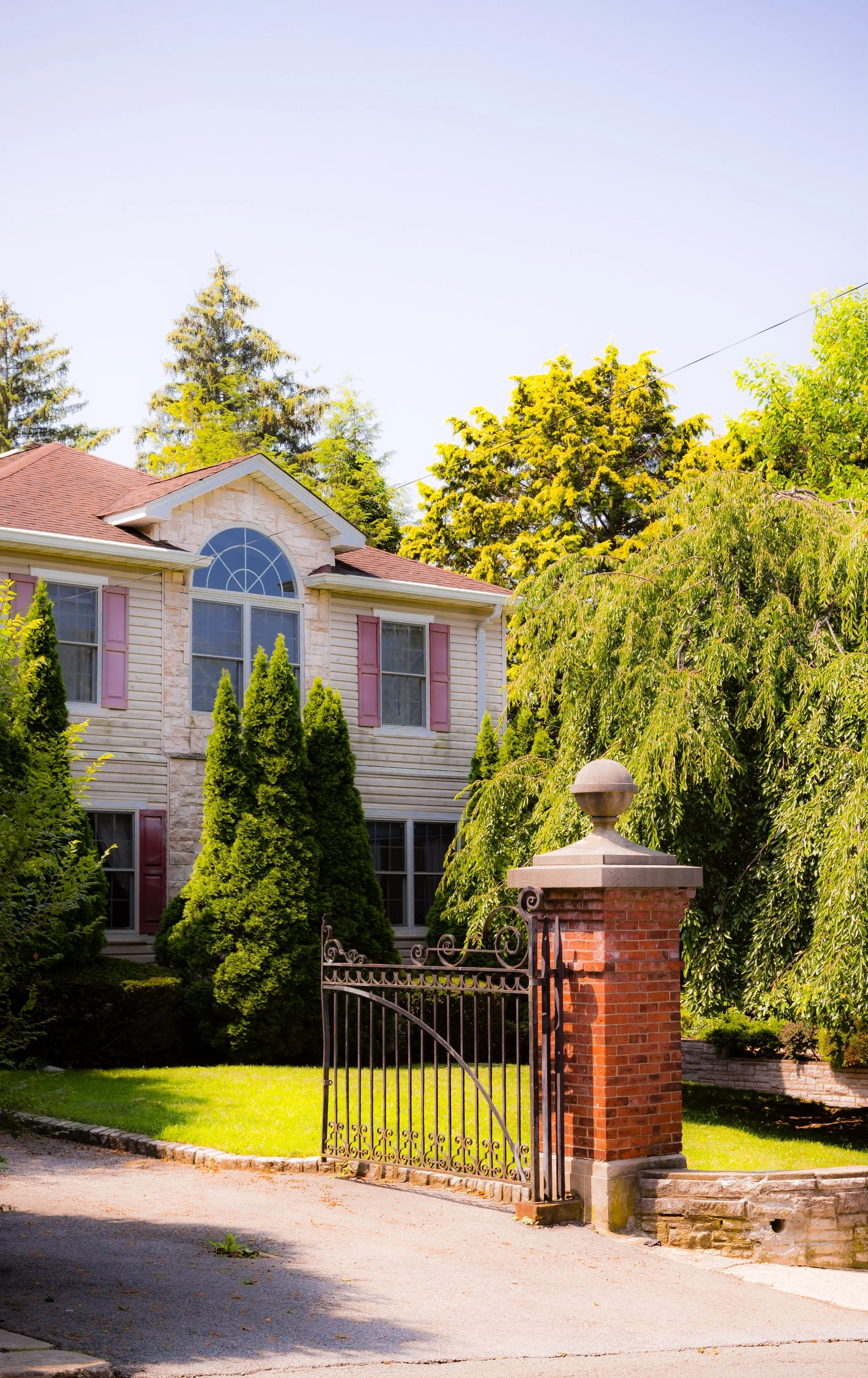 A two-story house with a brick and siding exterior, pink window shutters, and an arched window at the top, surrounded by green trees and a manicured lawn, with a black wrought iron gate at the driveway entrance.