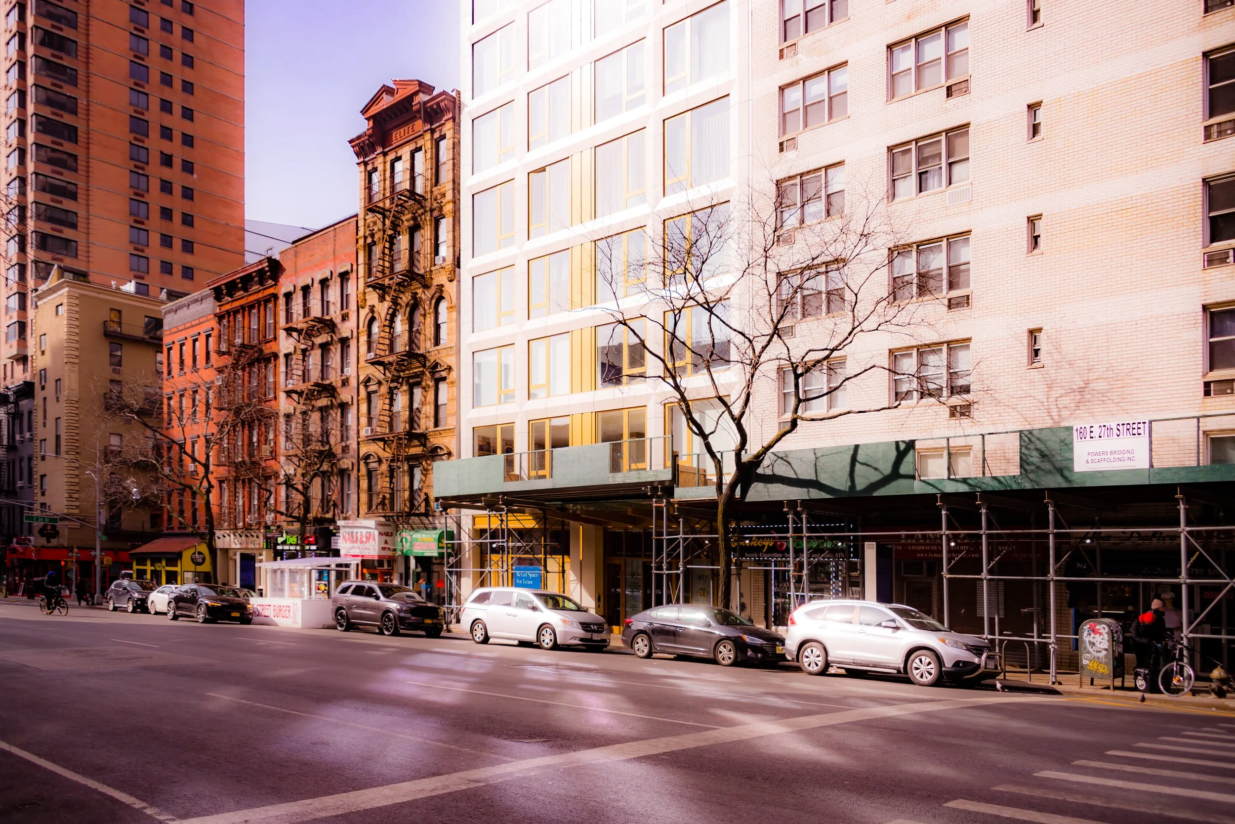 City street scene with buildings, parked cars, and a person on a bicycle. Trees are visible, and some buildings are under construction with scaffolding.