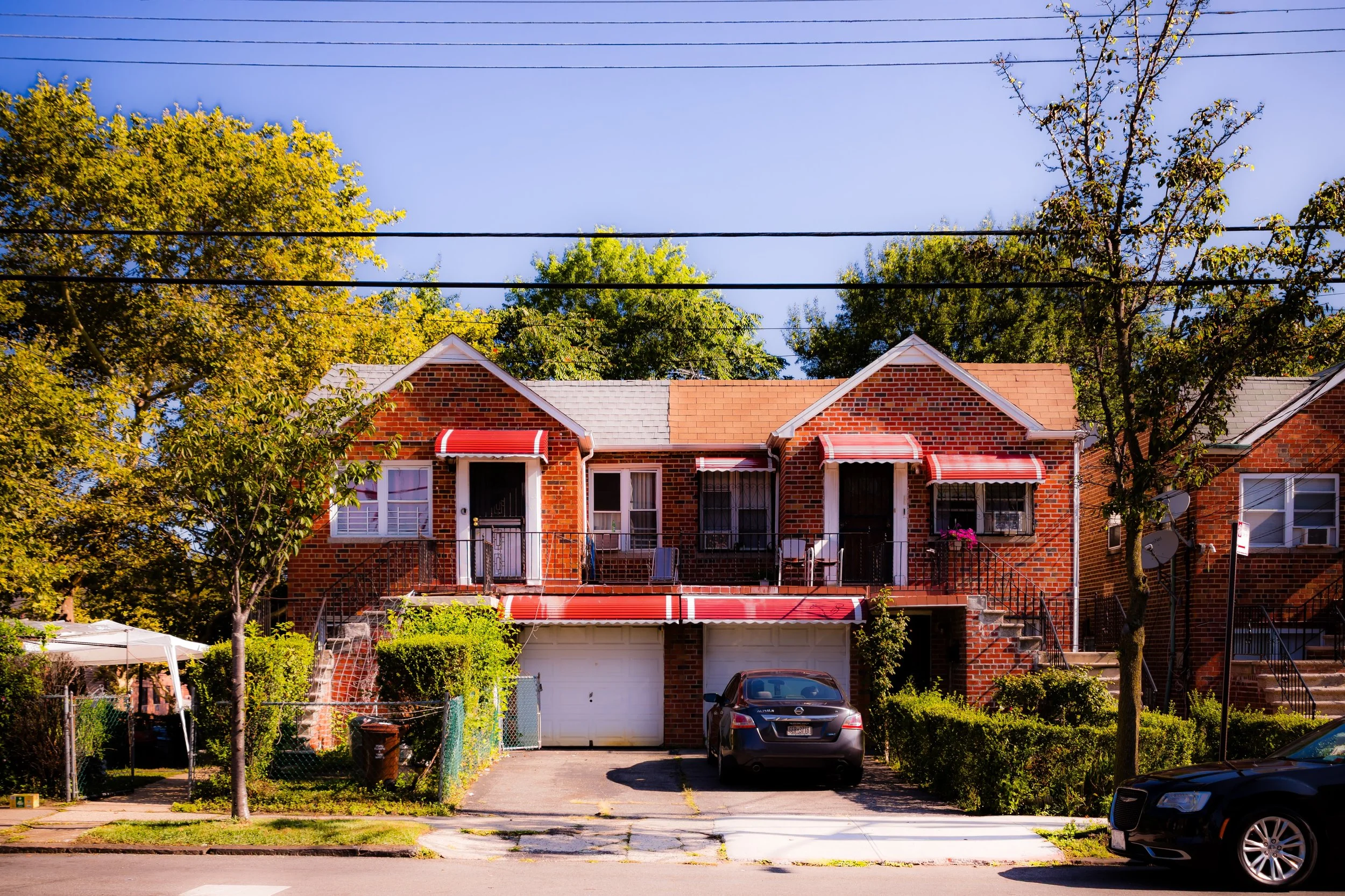 A row of brick townhouses with small front porches and staircases, cars parked in front, and trees on either side, under a clear blue sky.
