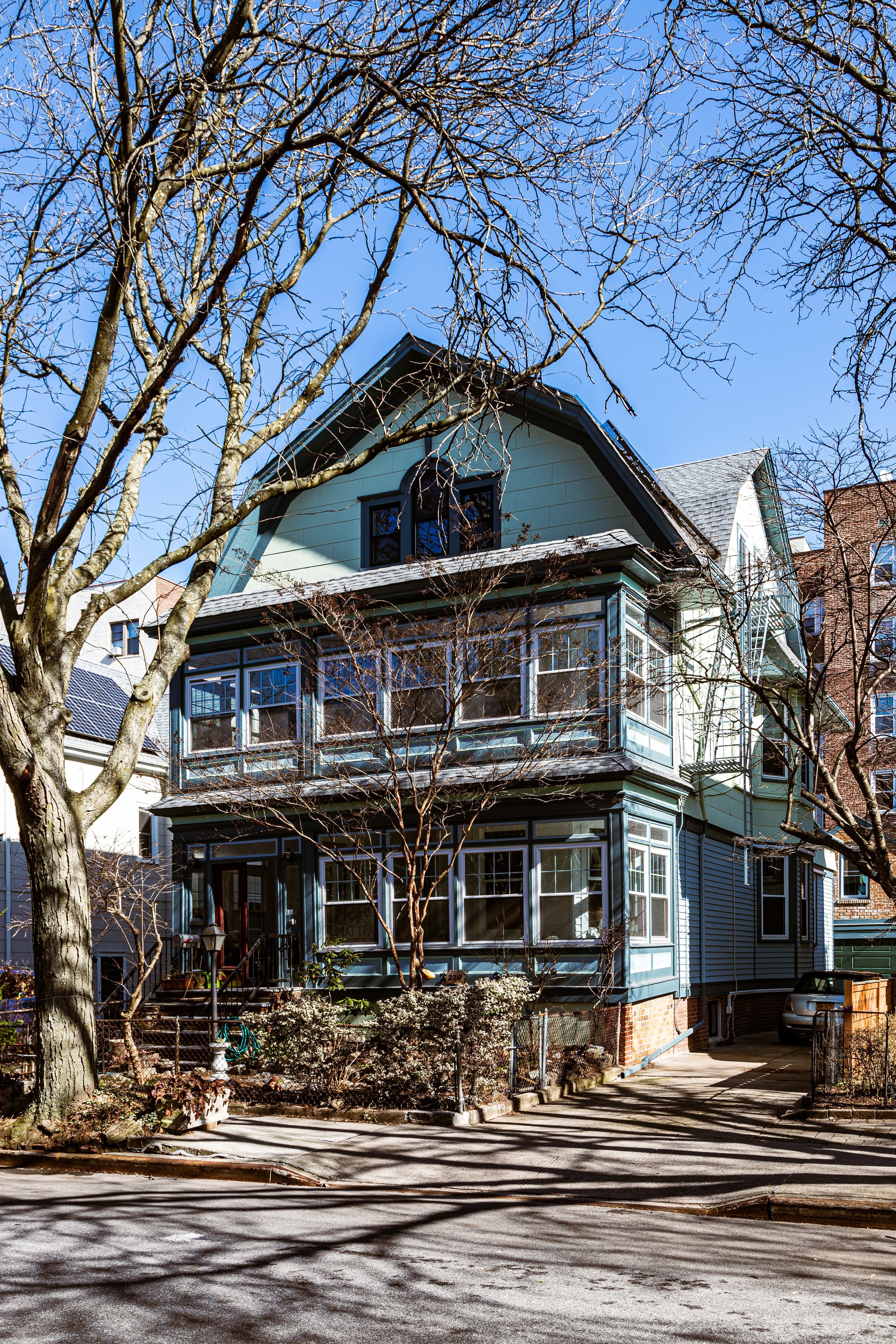 A three-story blue house with front porch, surrounded by a sidewalk, trees, and neighboring buildings under a clear blue sky.